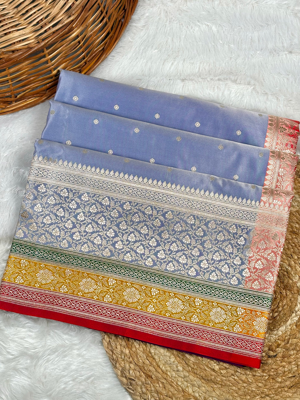 A folded saree with intricate floral patterns and borders, displayed on a white faux fur surface next to a woven wicker basket.