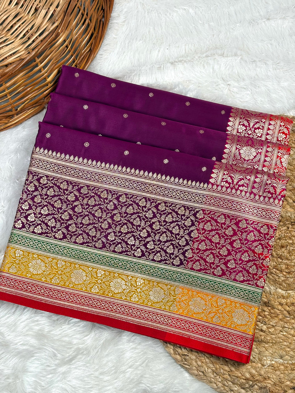 A folded purple Banarasi silk saree with intricate gold zari floral patterns and borders, displayed on a white faux fur surface with a wicker basket.