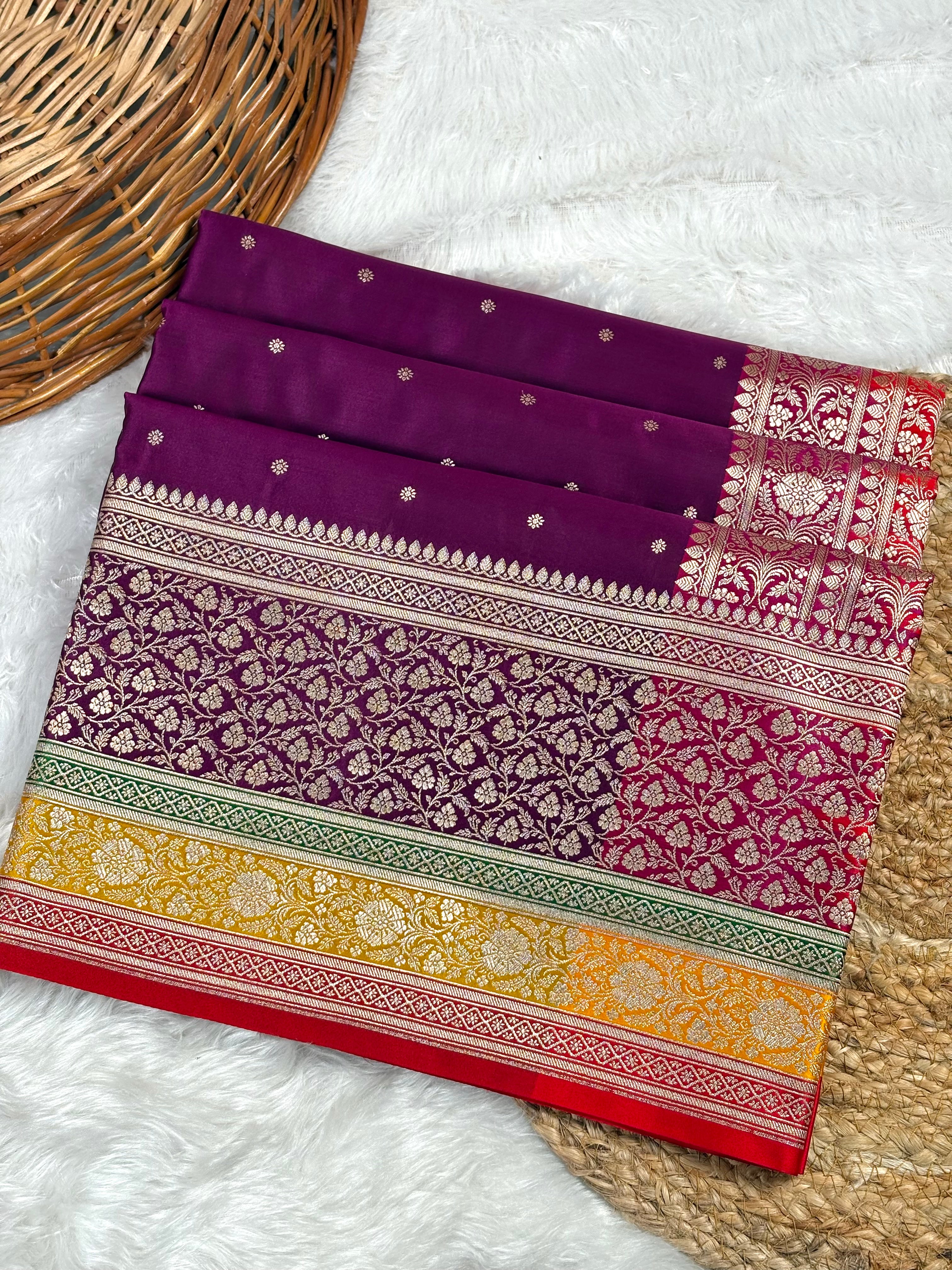 A folded purple Banarasi silk saree with intricate gold zari floral patterns and borders, displayed on a white faux fur surface with a wicker basket.