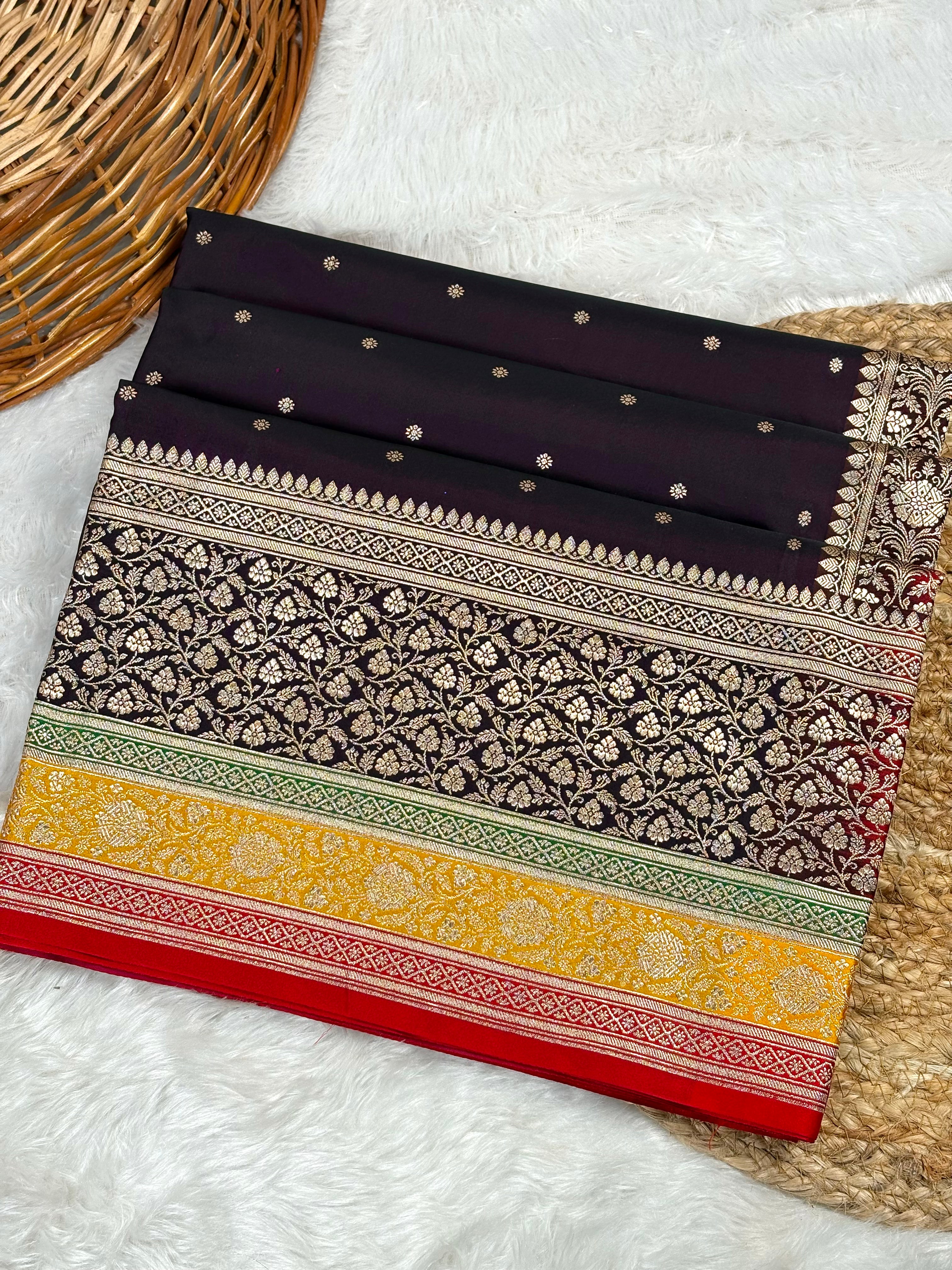 A folded Black Banarasi silk saree with intricate gold zari floral patterns, a yellow and green border, and a red bottom edge, displayed on a white faux fur rug next to a woven basket.