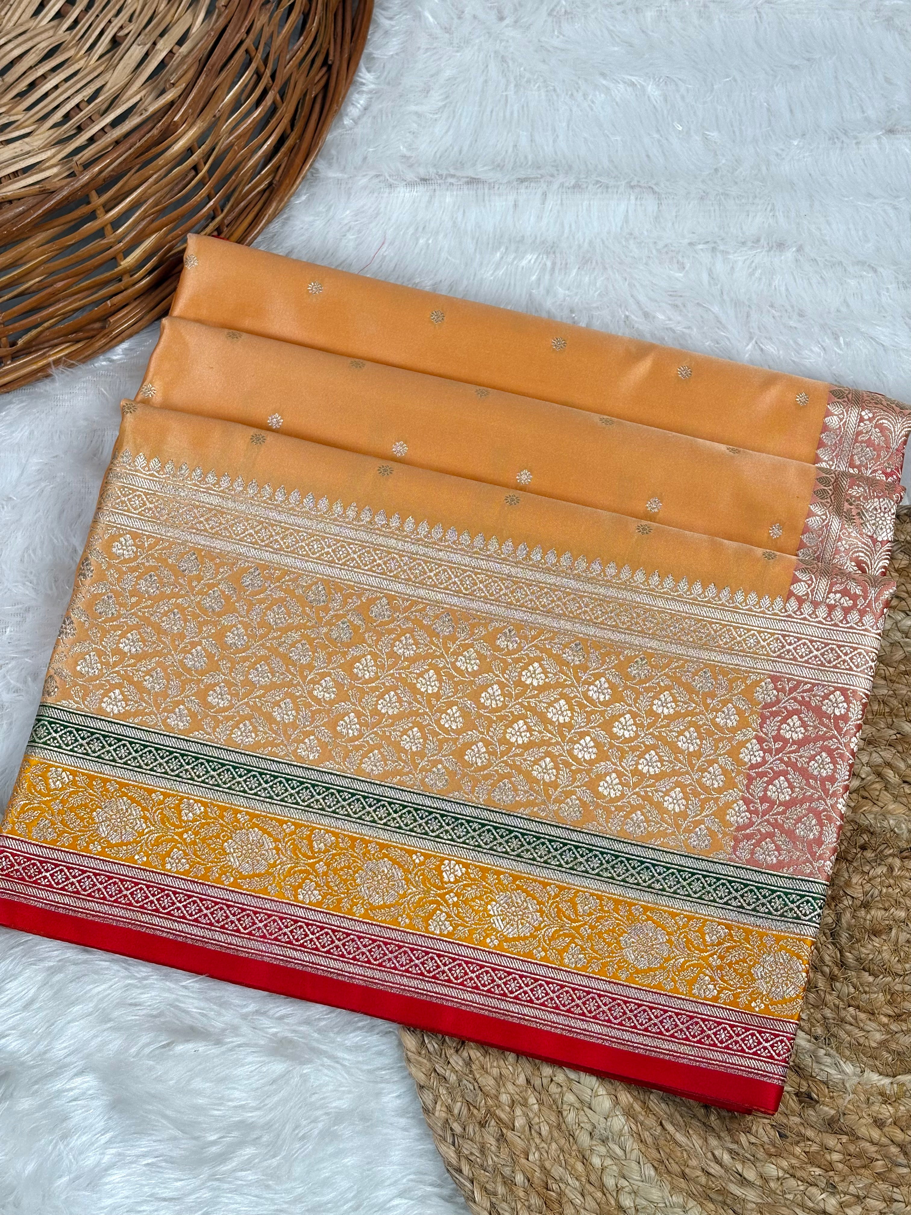 A folded peach-colored silk saree with intricate gold zari patterns, a red border, and green and gold decorative stripes, displayed on a white faux fur surface next to a woven wicker basket.