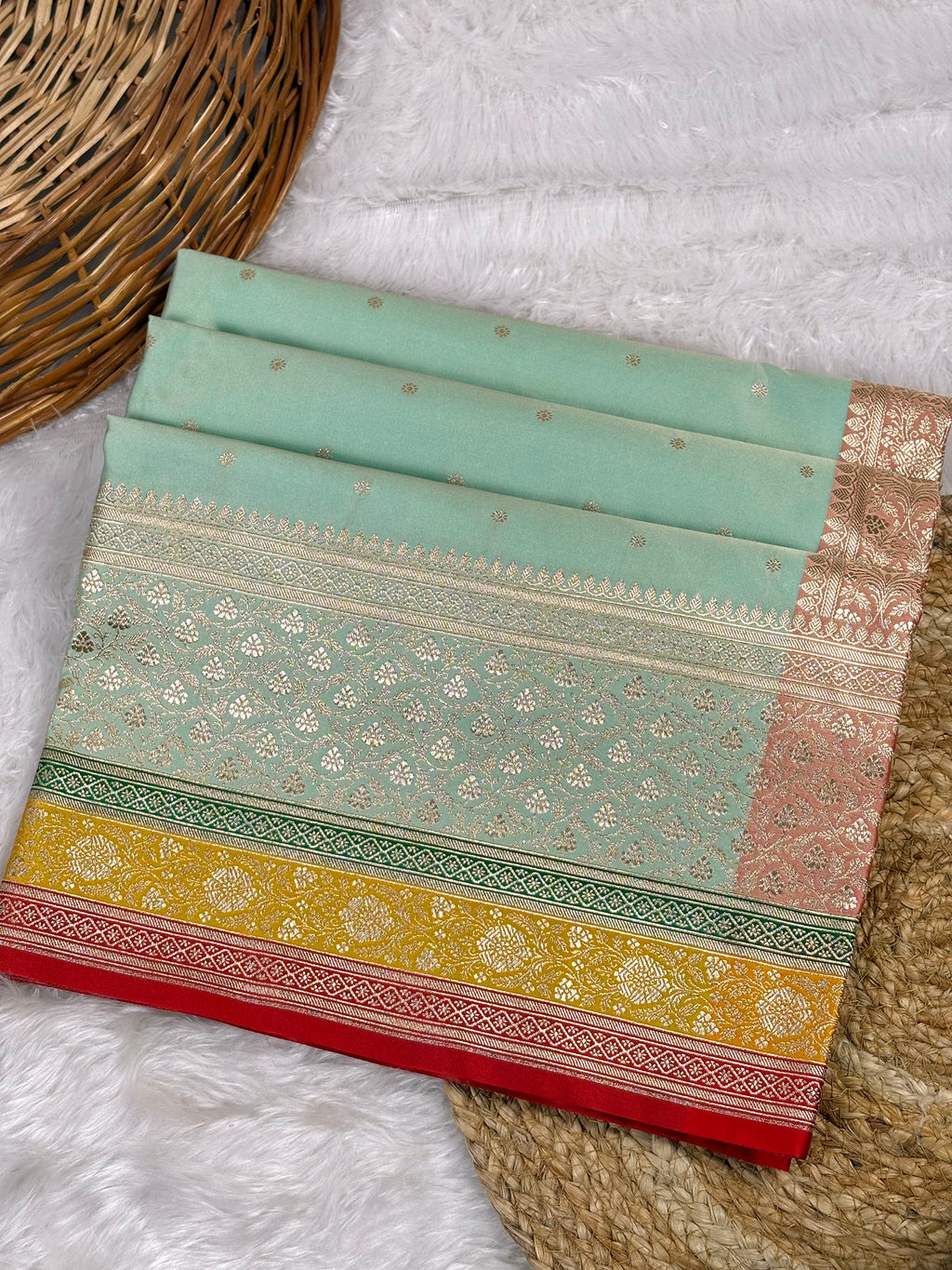 A folded mint green saree with intricate gold zari weaving, a yellow-gold and red border, displayed on a white faux fur surface next to a woven wicker basket.