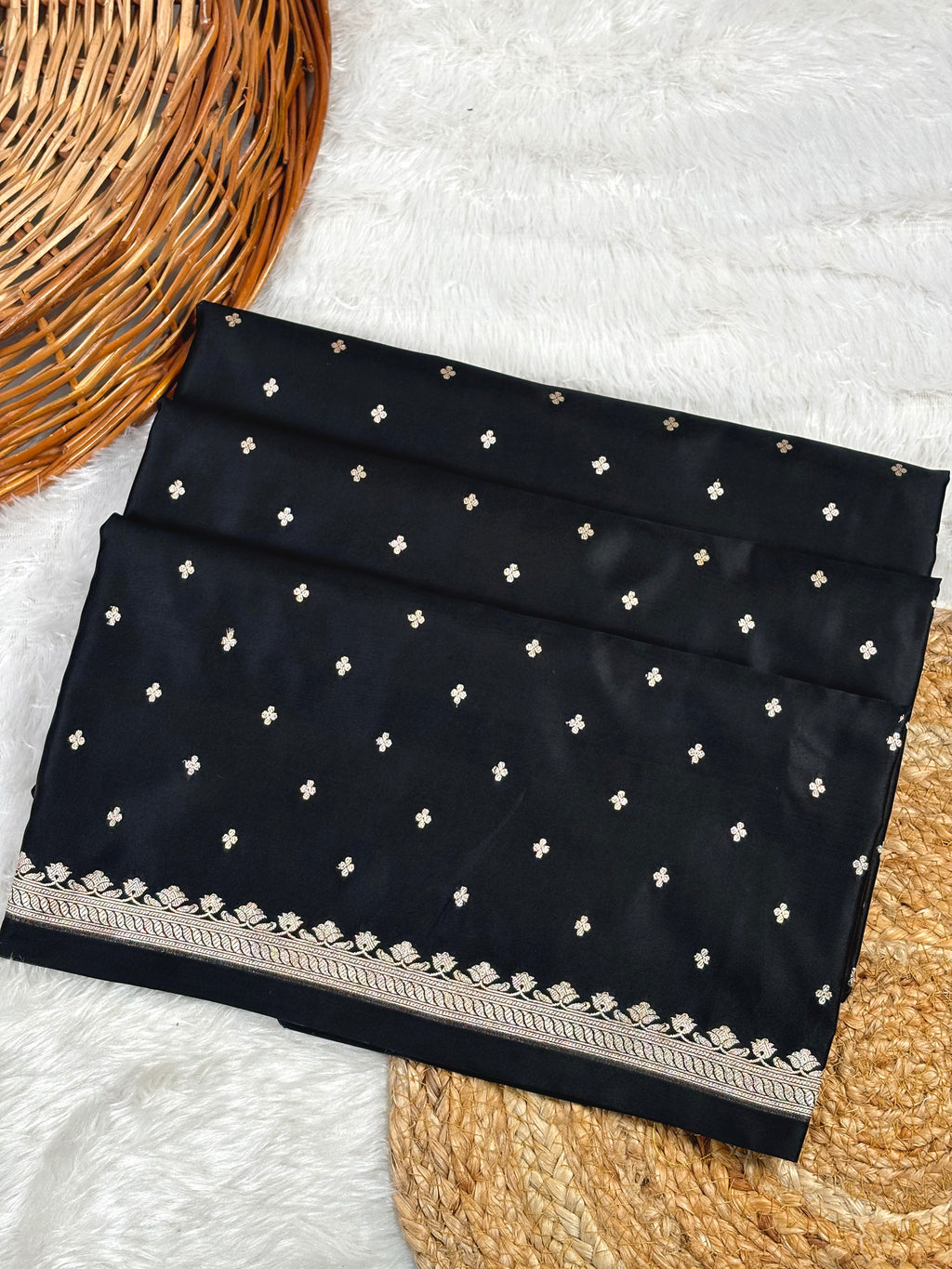 mashru katan black saree, decorated with small gold buti motifs and a detailed gold border, displayed on a white faux fur surface with a wicker basket.