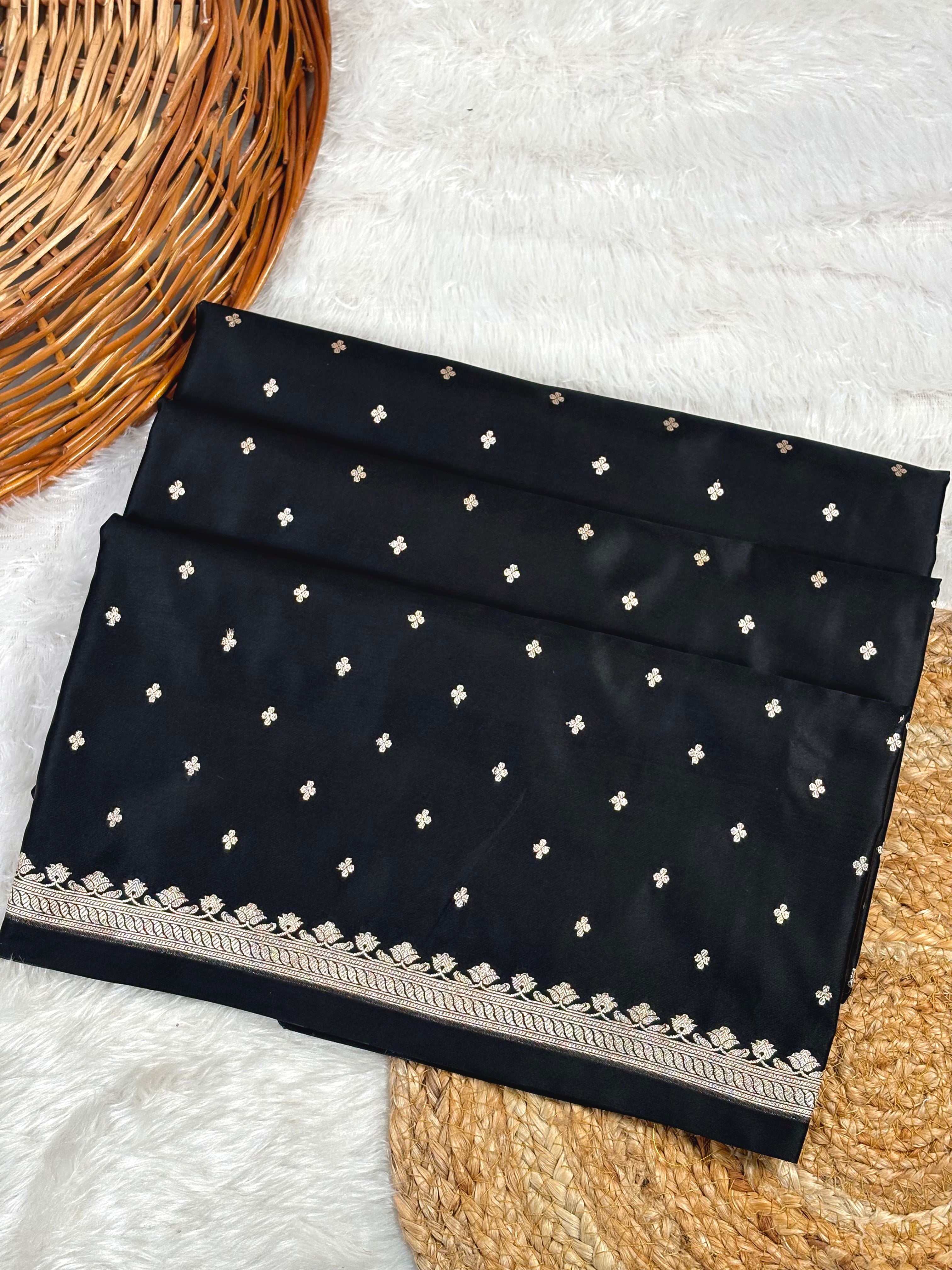 mashru katan black saree, decorated with small gold buti motifs and a detailed gold border, displayed on a white faux fur surface with a wicker basket.