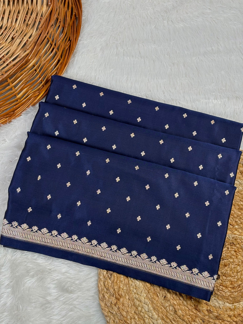 A folded navy blue saree, decorated with small white buti motifs and a detailed floral border, displayed on a white faux fur surface with a wicker basket.