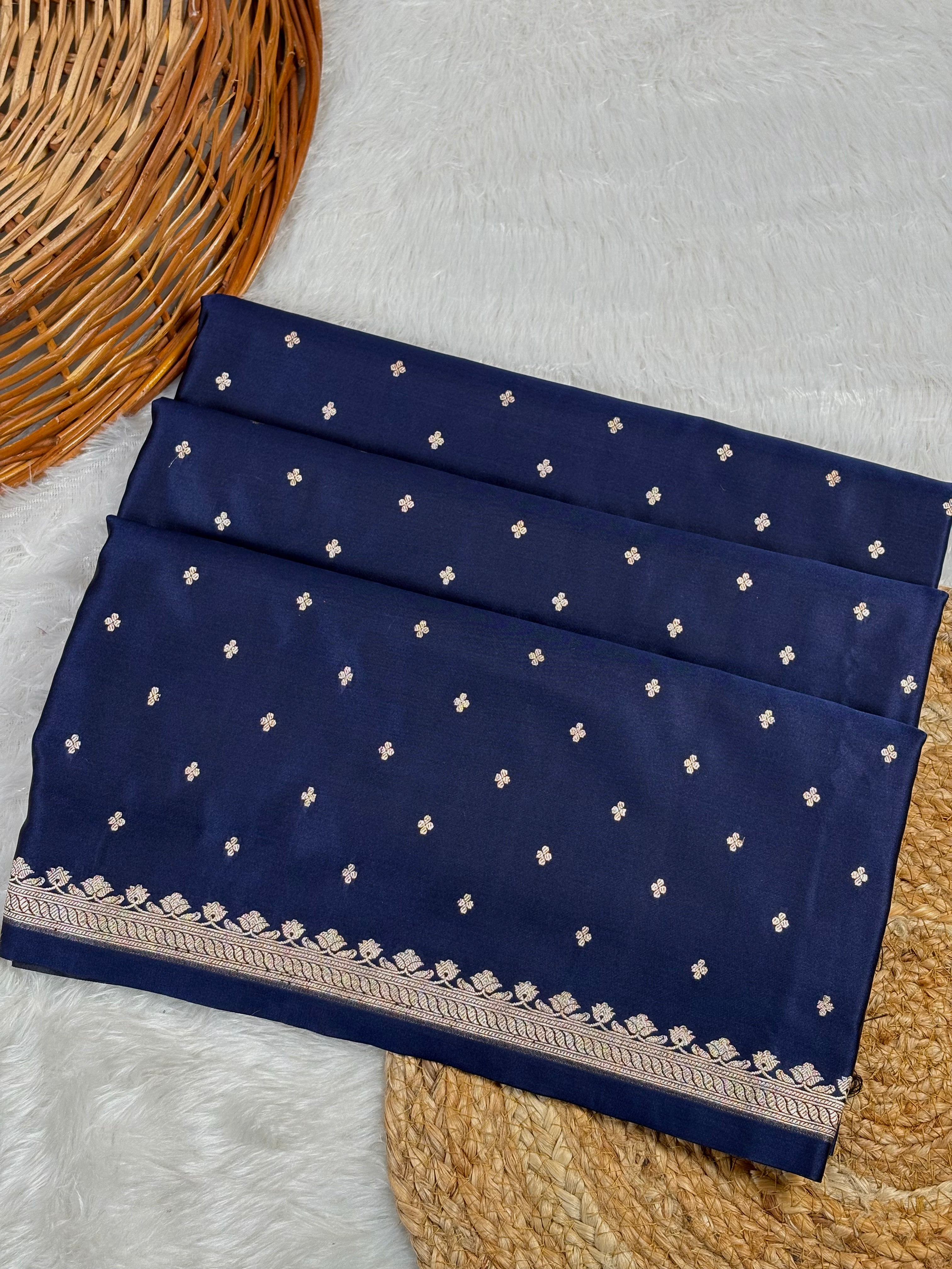 A folded navy blue saree, decorated with small white buti motifs and a detailed floral border, displayed on a white faux fur surface with a wicker basket.