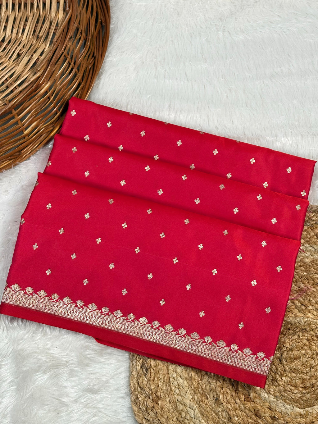 A folded red mashru katan saree decorated with small gold motifs and a gold border, displayed on a white faux fur surface with a wicker basket.