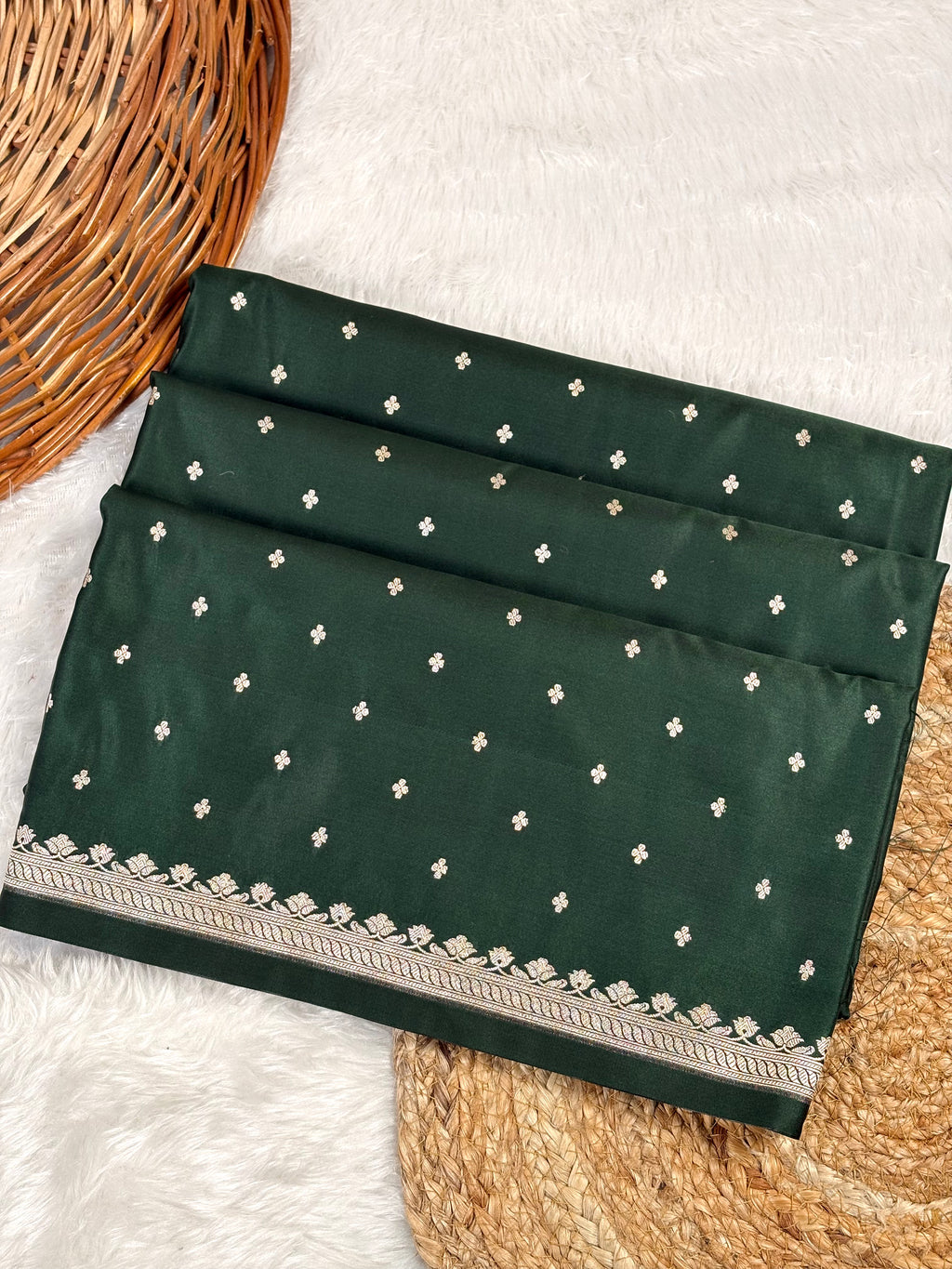 A folded bottle green saree, decorated with small gold motifs and a detailed gold border, displayed on a white faux fur surface with a wicker basket.