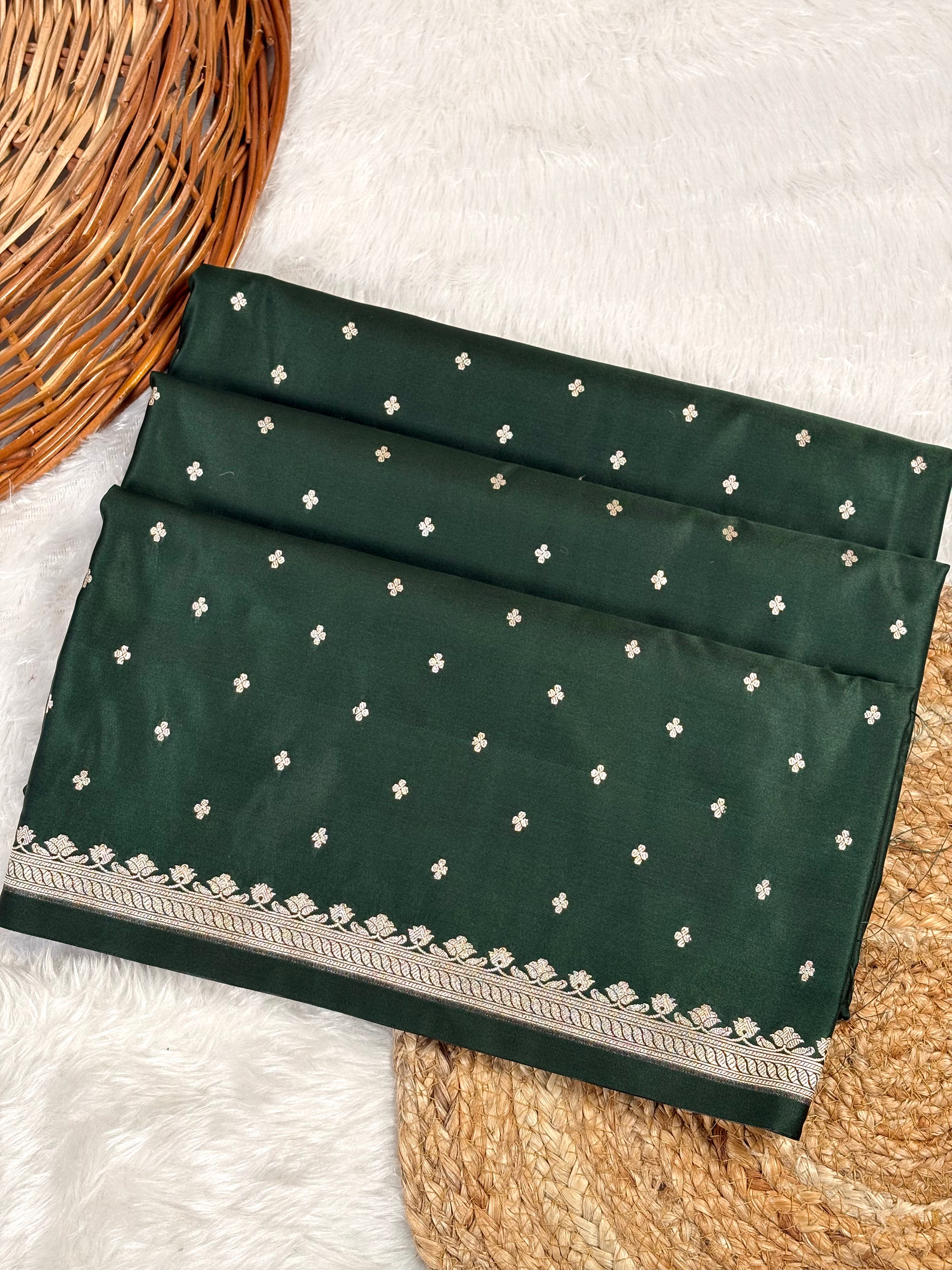 A folded bottle green saree, decorated with small gold motifs and a detailed gold border, displayed on a white faux fur surface with a wicker basket.