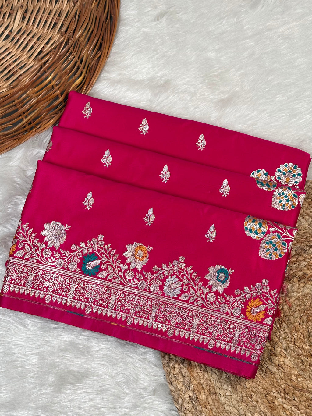 A folded red pink Banarasi silk saree with intricate sona rupa zari weaving, floral motifs, and a detailed border, displayed on a white faux fur rug next to a woven wicker basket.