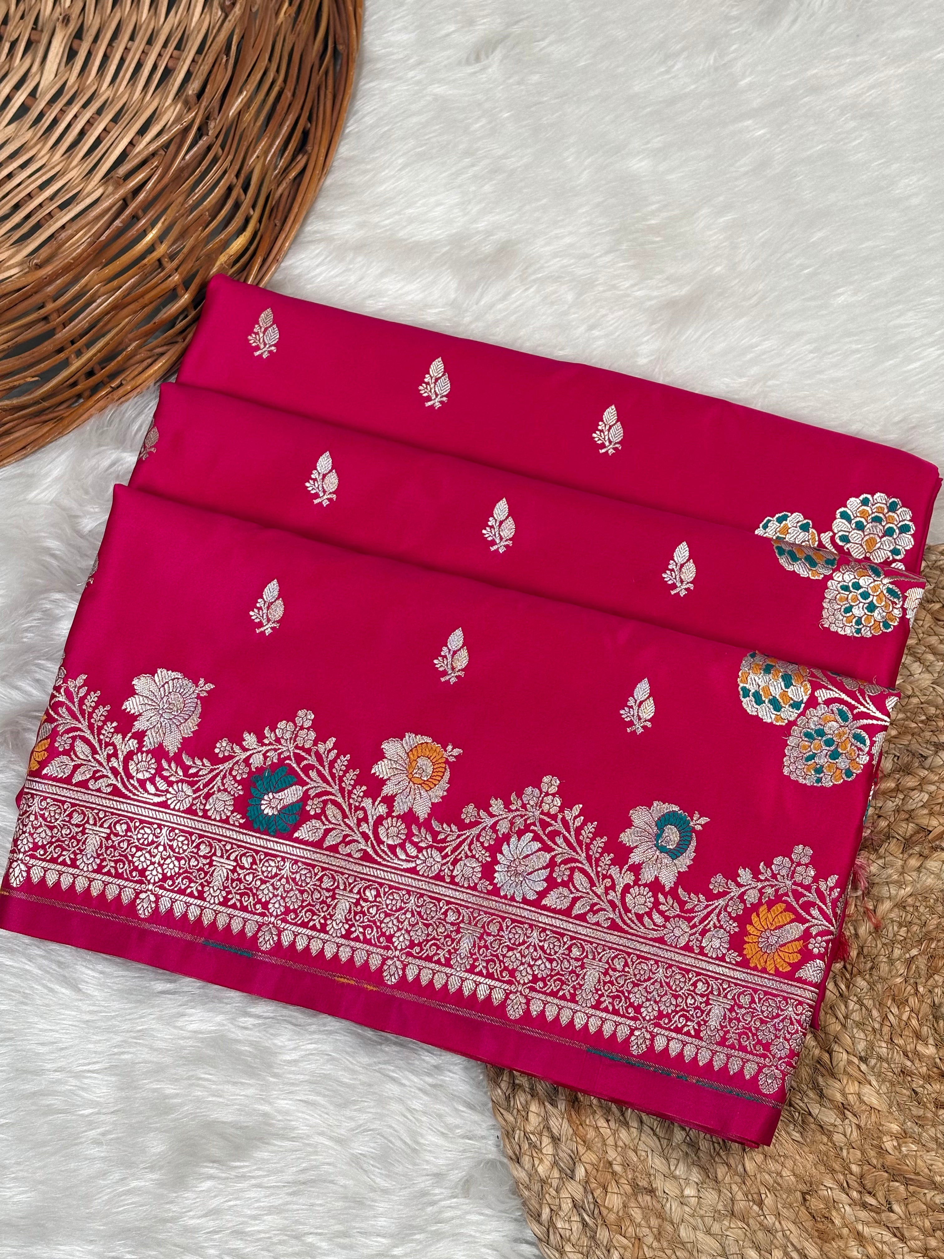 A folded red pink Banarasi silk saree with intricate sona rupa zari weaving, floral motifs, and a detailed border, displayed on a white faux fur rug next to a woven wicker basket.