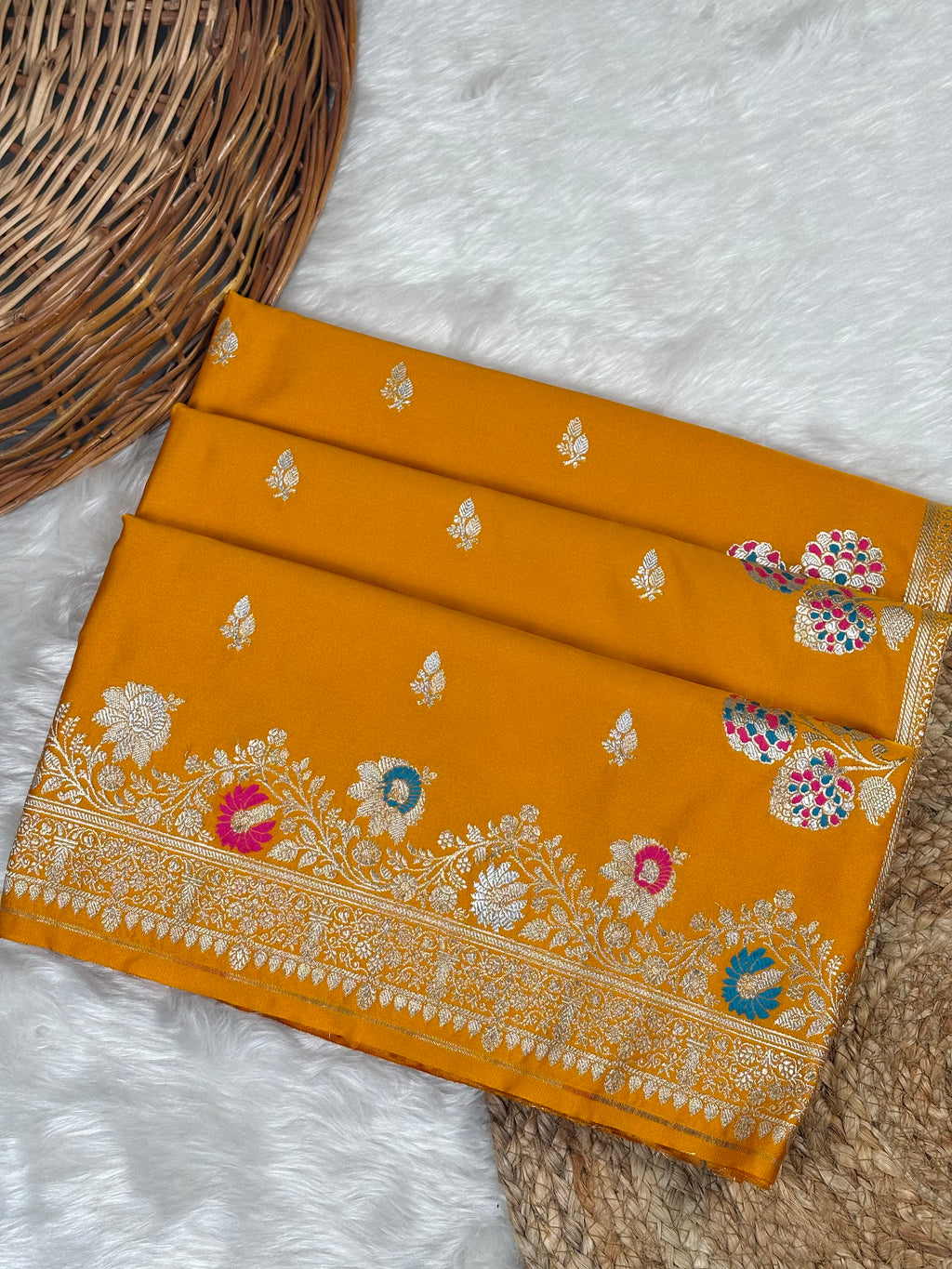 A folded mustard yellow silk saree with intricate sona rupa zari weaving and colorful floral motifs, displayed on a white faux fur surface next to a woven basket.