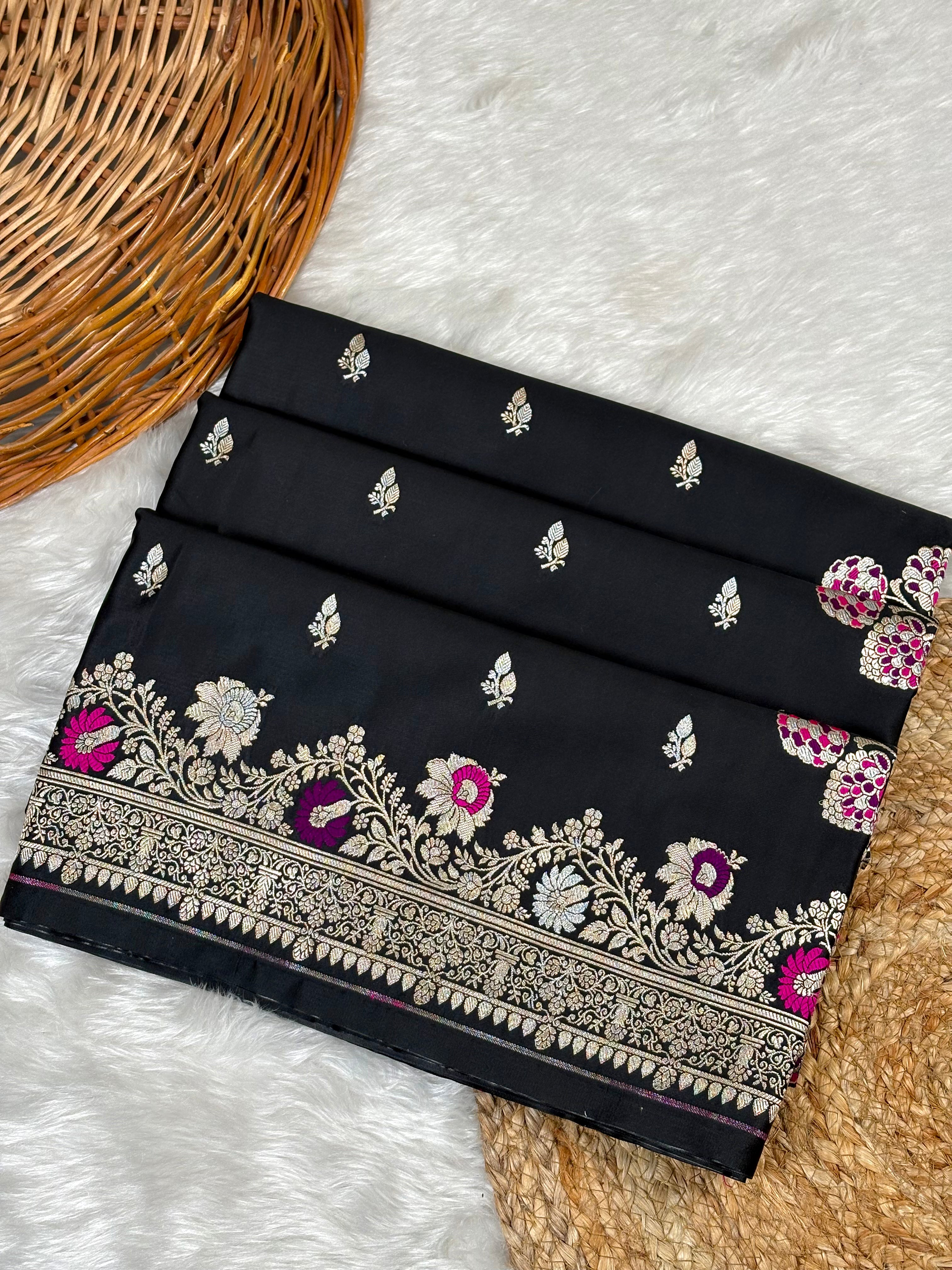 A folded black Banarasi silk saree with intricate sona rupa zari weaving and floral motifs, displayed on a white faux fur rug next to a woven wicker basket.