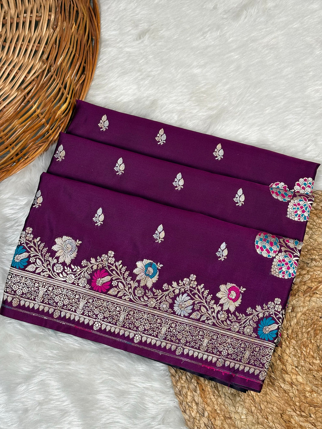 A folded deep purple silk saree with intricate rose gold zari weaving, floral motifs, and a detailed border, displayed on a white faux fur surface next to a wicker basket.
