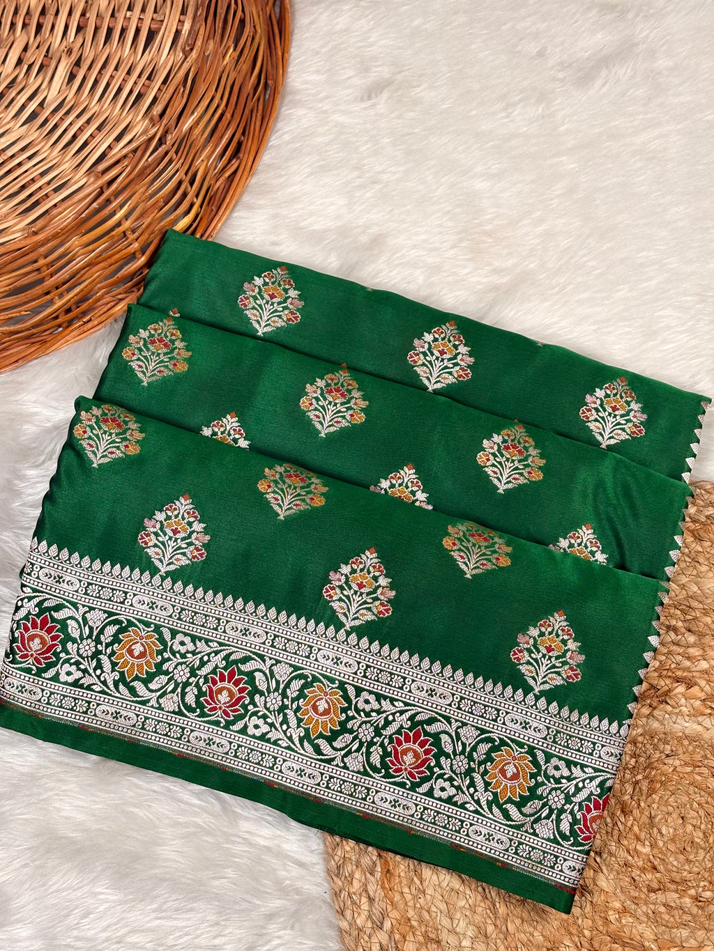 A folded dark green saree with floral  buta and a wide decorative border, styled on a white faux-fur surface with a wicker basket.