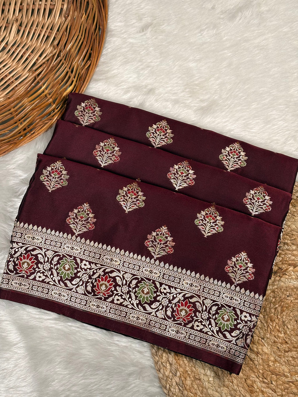 A folded deep maroon saree with gold floral motifs and a detailed gold border, displayed on a white faux fur surface next to a wicker basket.