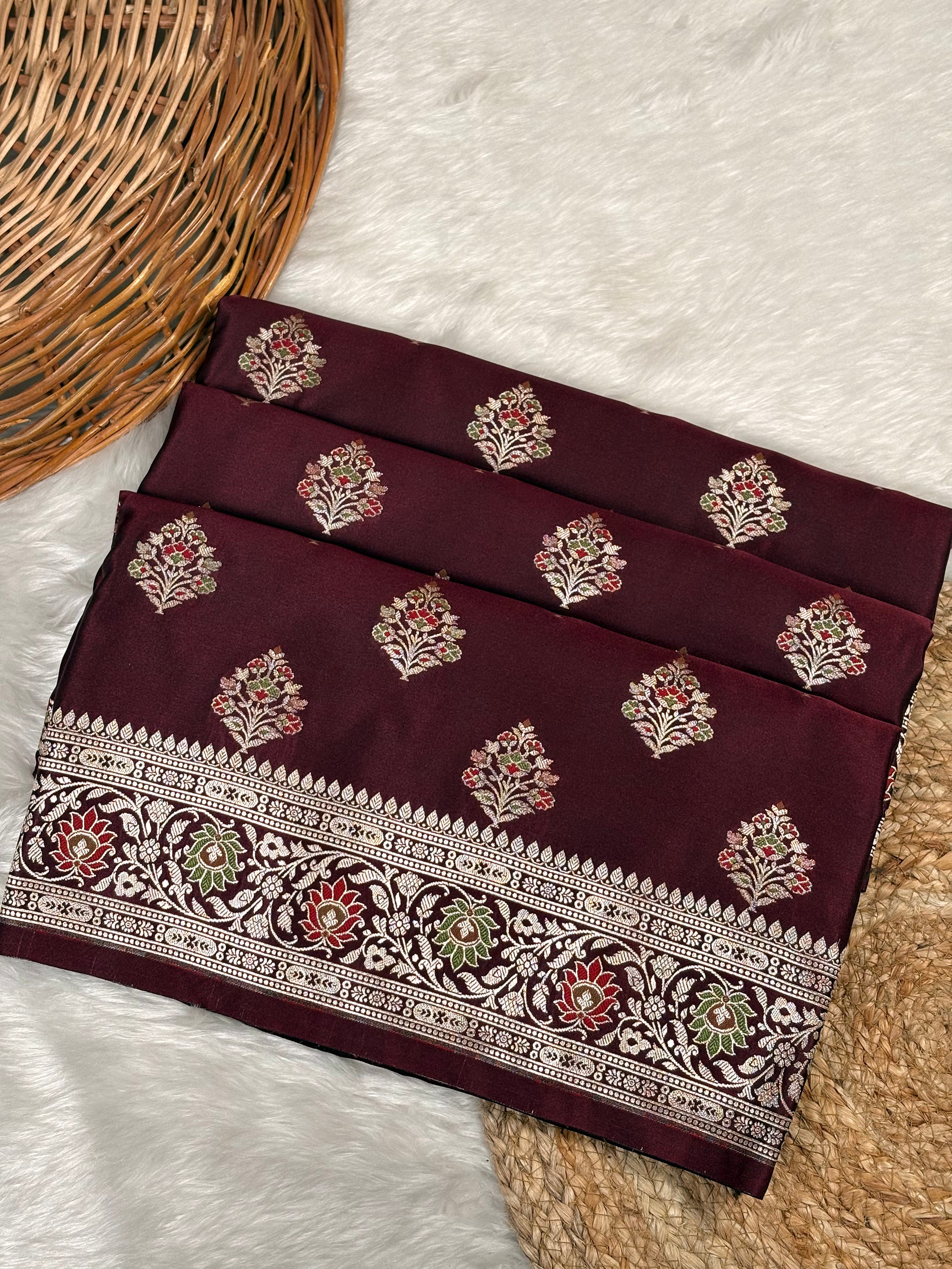 A folded deep maroon saree with gold floral motifs and a detailed gold border, displayed on a white faux fur surface next to a wicker basket.