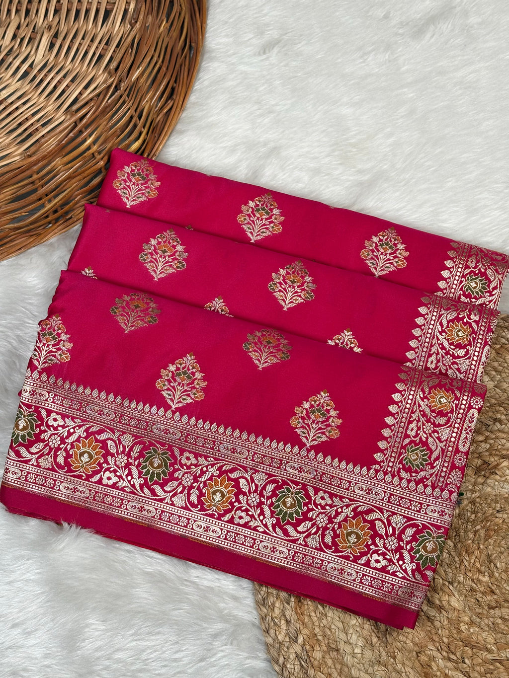 A folded red-pink saree with intricate gold and green mina in a floral woven buta and a wide decorative border, displayed on a white faux fur surface next to a woven wicker basket