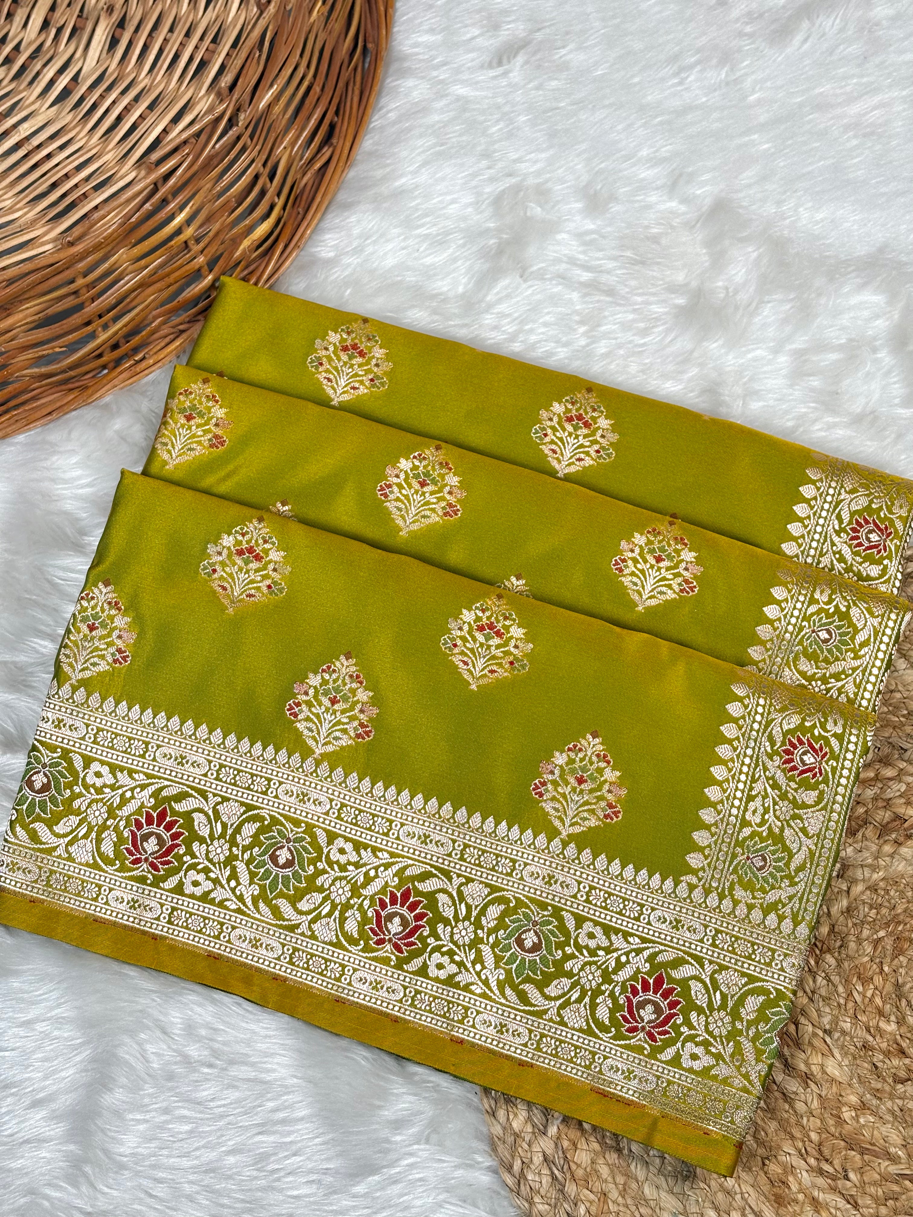 An olive green Banarasi silk saree with intricate gold zari weaving and floral motifs, displayed on a white faux fur surface next to a wicker basket.