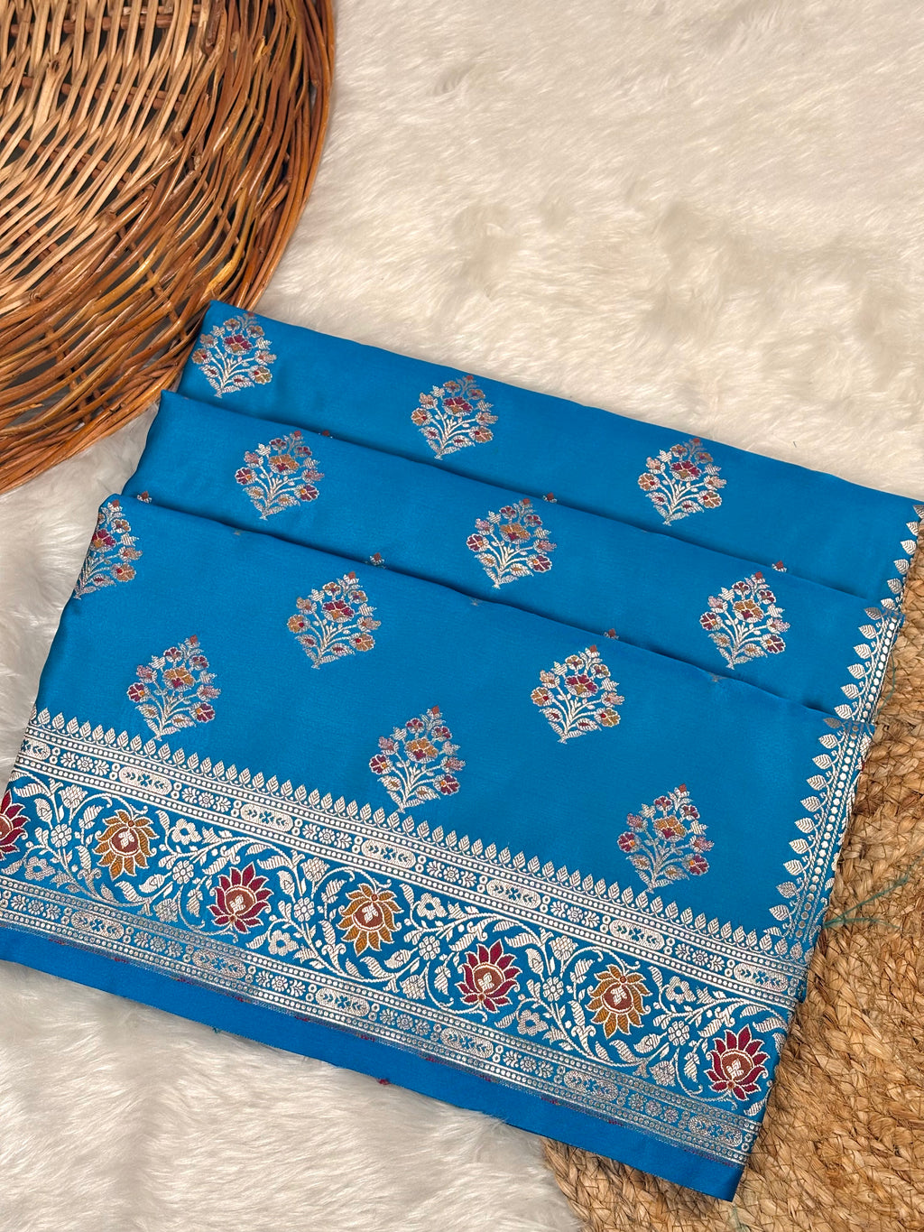 A folded blue saree with intricate floral buta and a detailed border, displayed on a white faux fur surface next to a wicker basket.