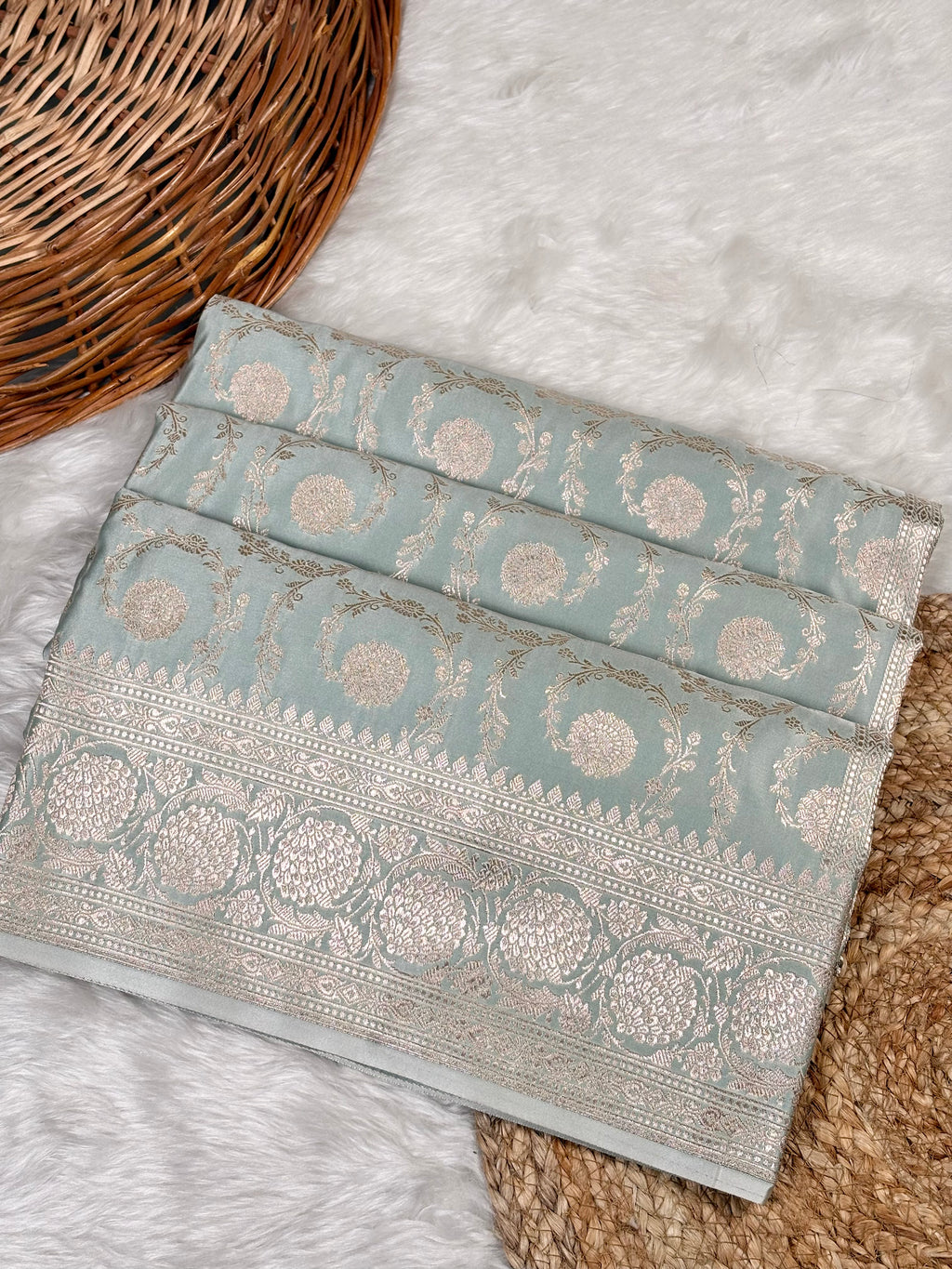 A folded light blue-green saree with intricate silver and gold zari floral motifs and borders, displayed on a white faux fur rug next to a wicker basket.