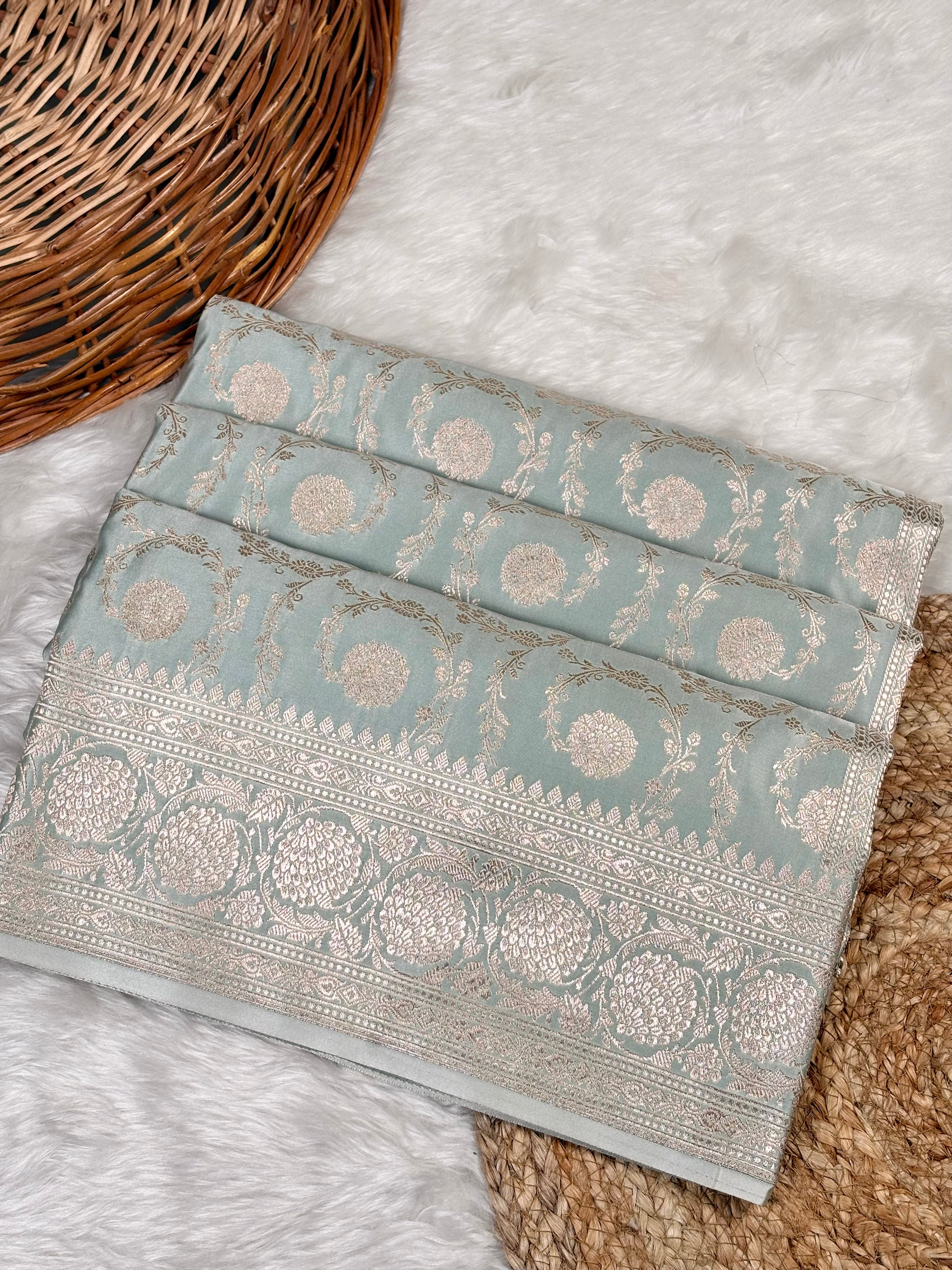 A folded light blue-green saree with intricate silver and gold zari floral motifs and borders, displayed on a white faux fur rug next to a wicker basket.