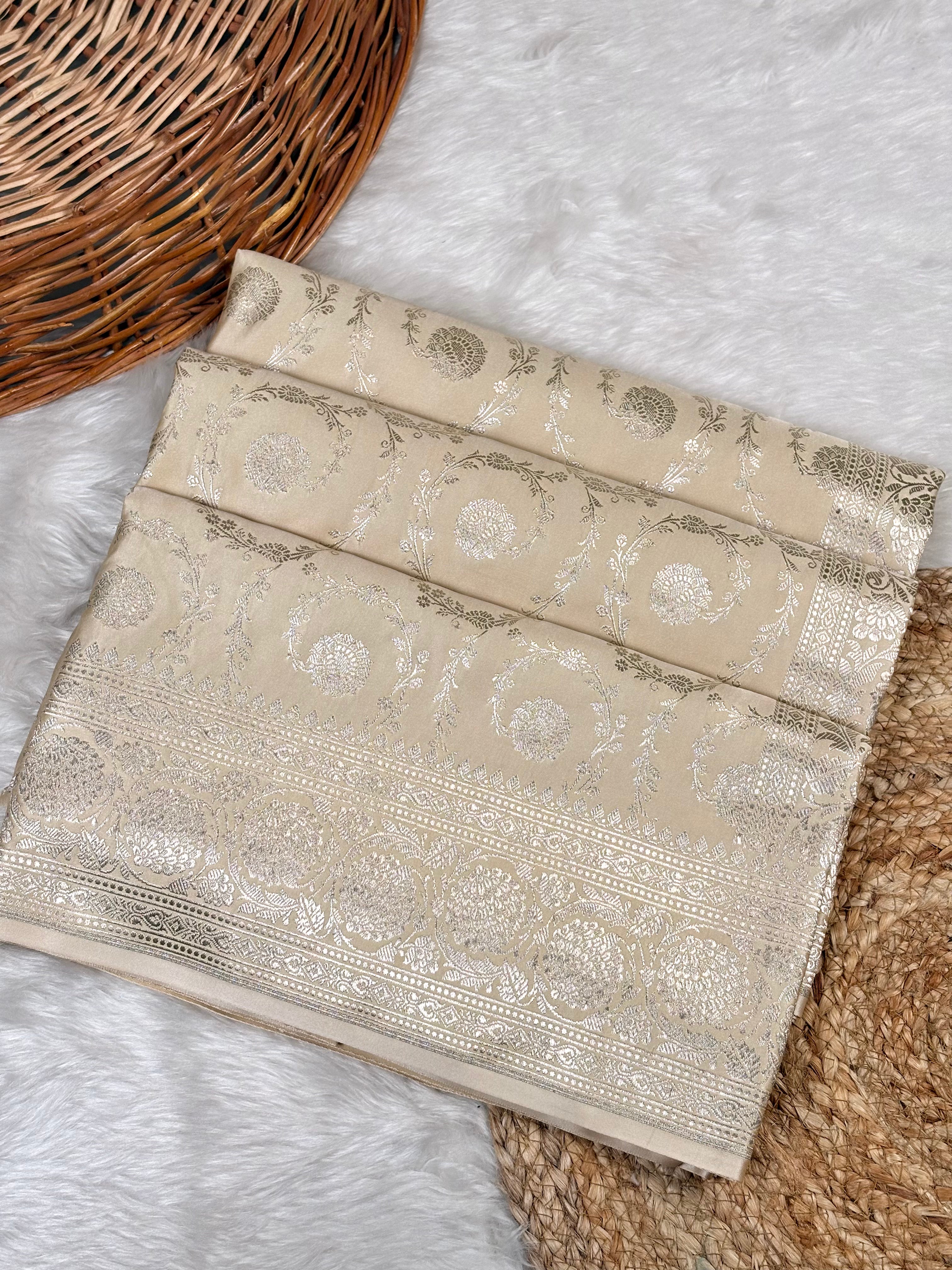A folded beige saree with ornate metallic silver woven patterns, displayed on a white faux fur surface next to a wicker basket.
