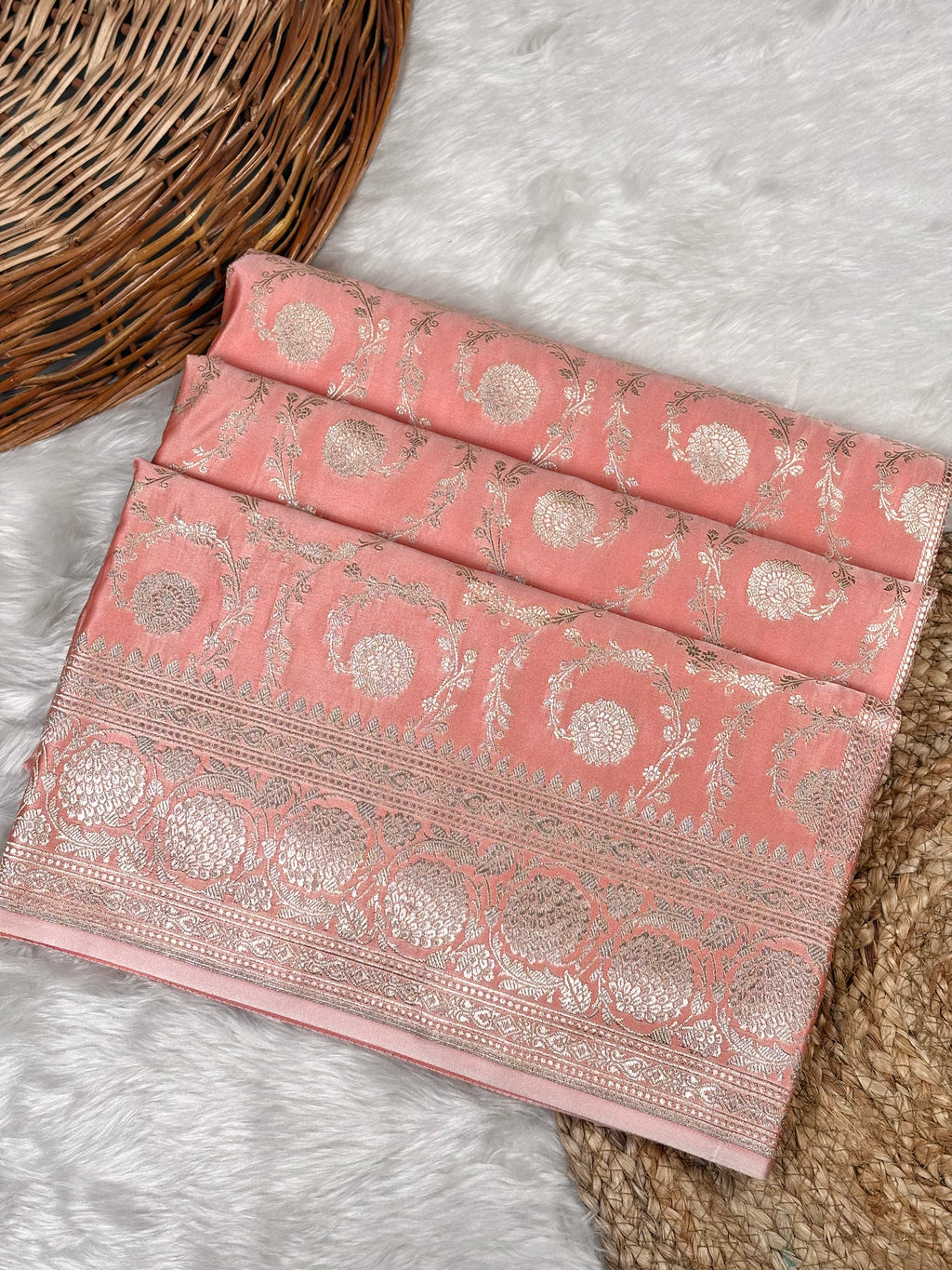 A folded pink saree with ornate metallic silver floral and paisley motifs, displayed on a white faux fur surface next to a woven wicker basket.