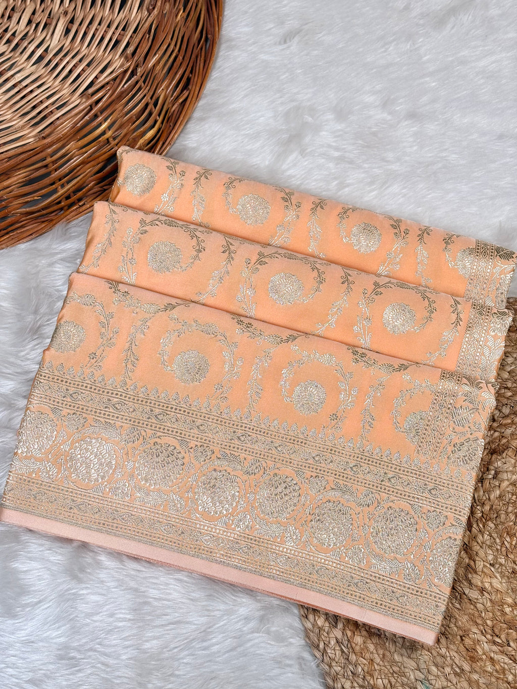 A peach-colored silk saree with intricate rose gold zari brocade patterns, folded neatly on a white faux fur rug next to a wicker basket.