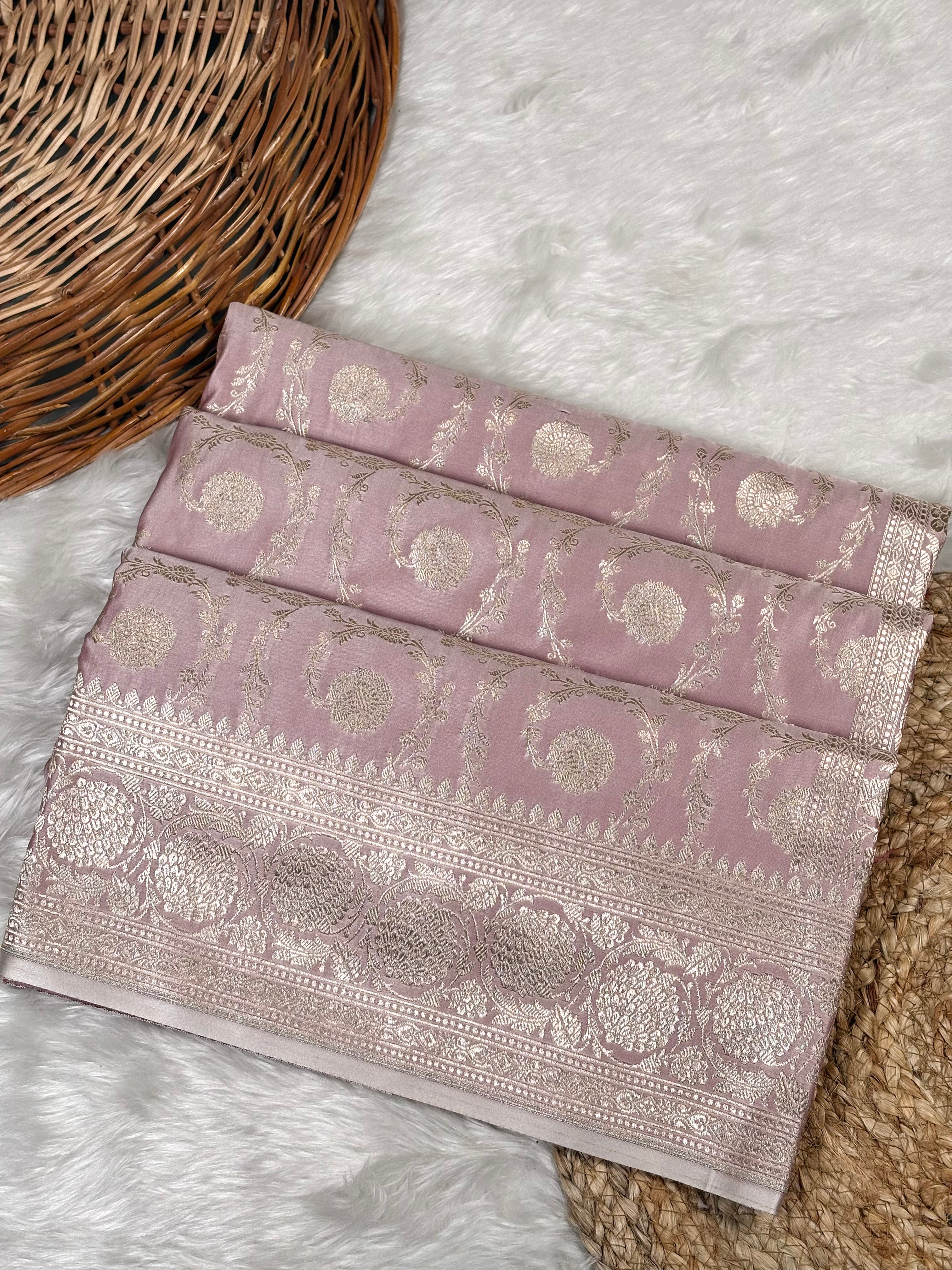 A folded dusty pink Banarasi silk saree with intricate silver zari floral motifs and borders, displayed on a white faux fur rug next to a wicker basket.