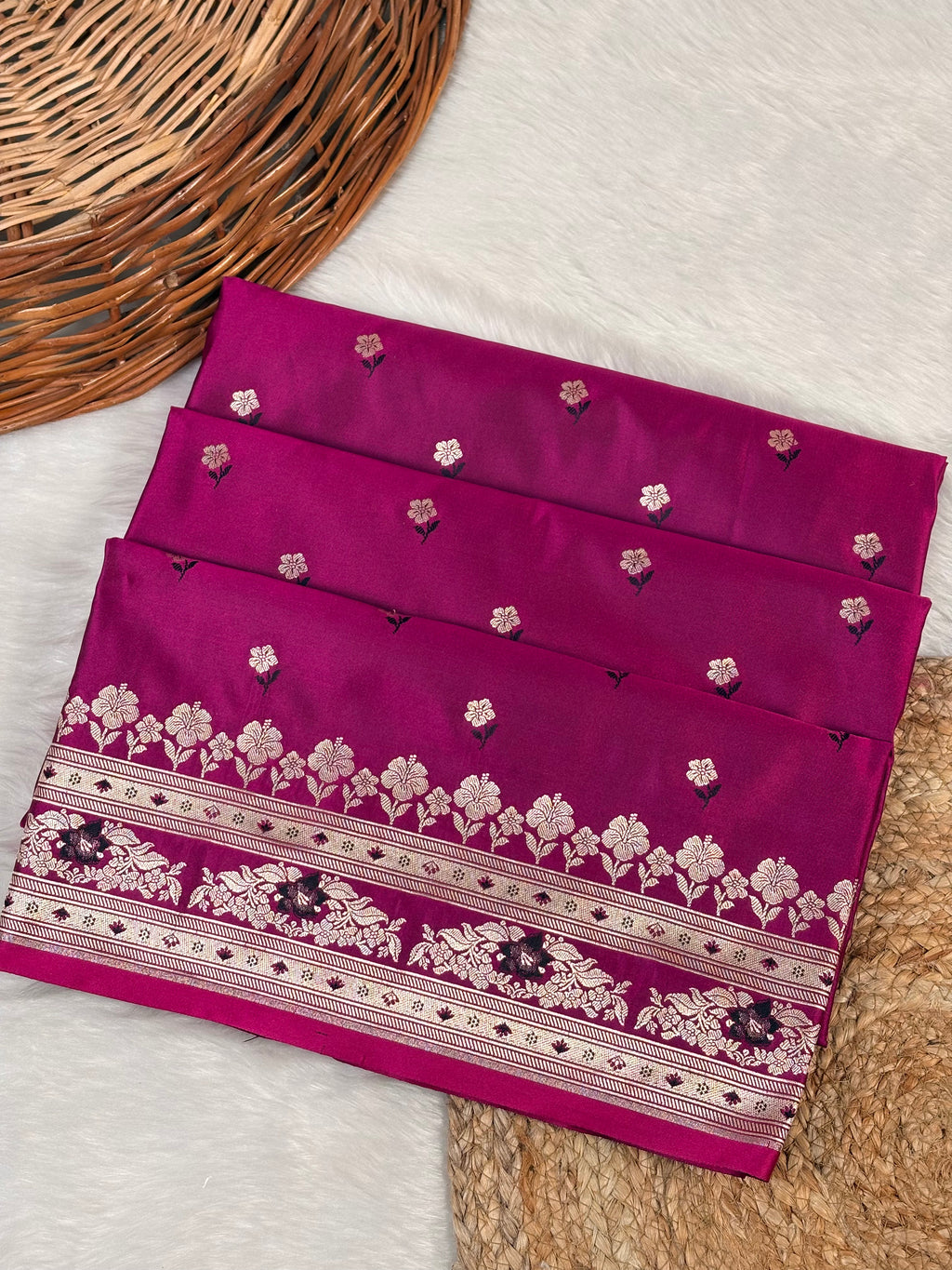 A folded purple saree with intricate rose gold zari weaving and a decorative border, displayed on a white faux fur surface next to a wicker mat.