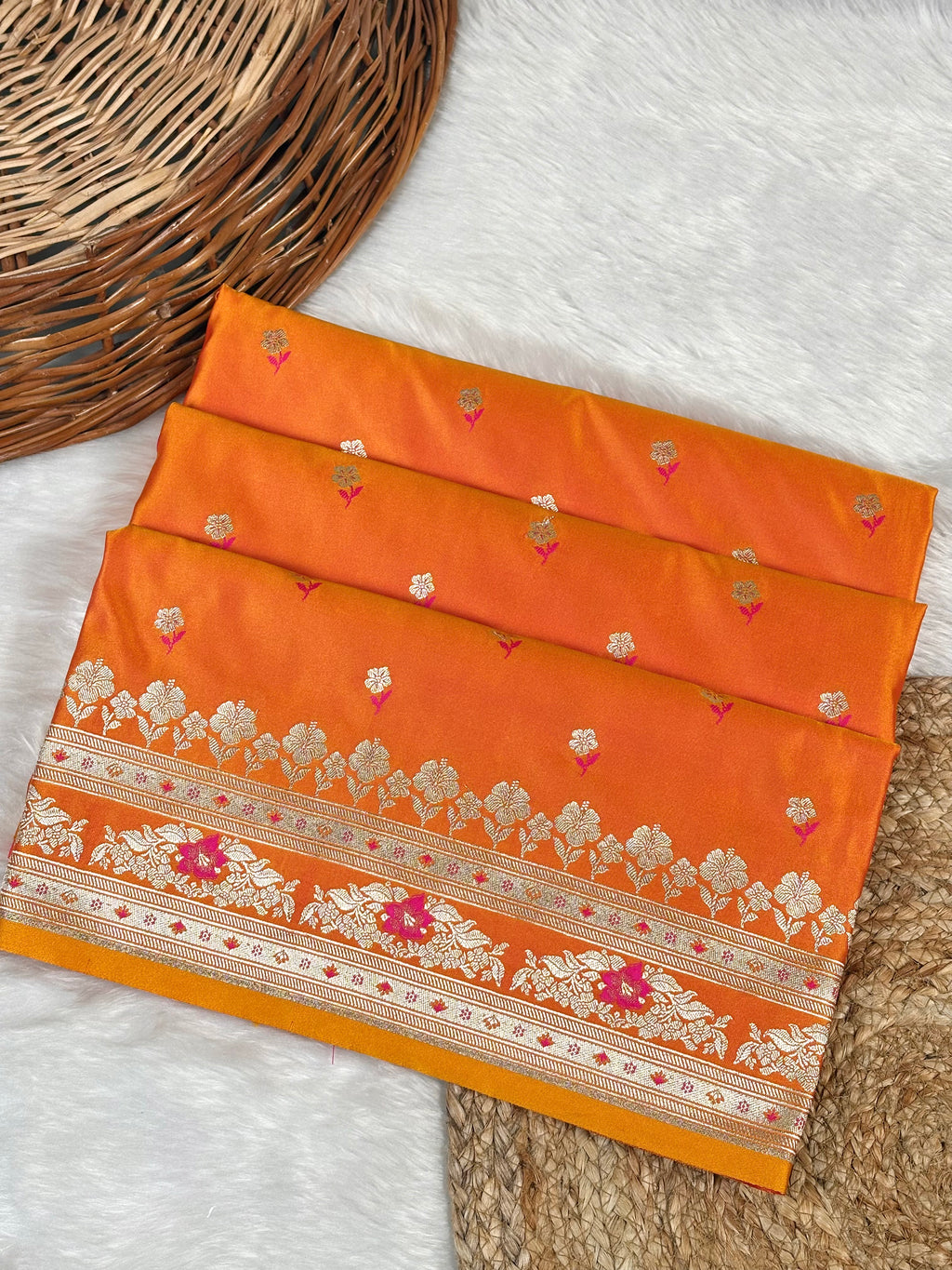An orange silk saree with intricate rose gold floral weaving and pink mina border, displayed in a folded stack on a white faux fur surface next to a wicker basket.