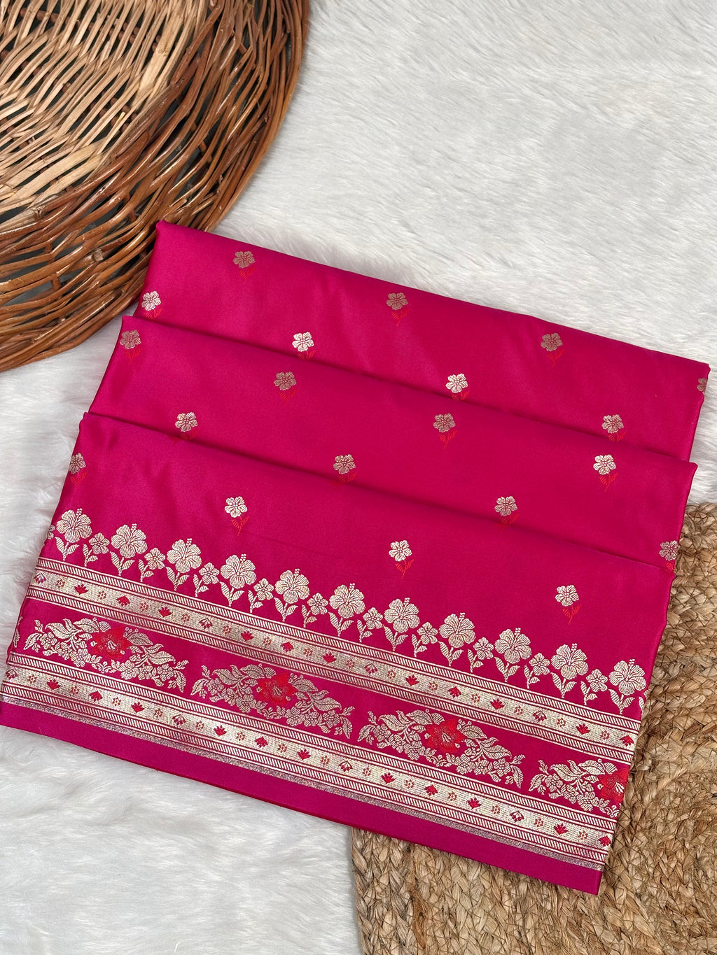 A folded magenta pink saree with intricate rose gold floral Banarasi zari weaving, displayed on a white faux fur surface next to a wicker basket.