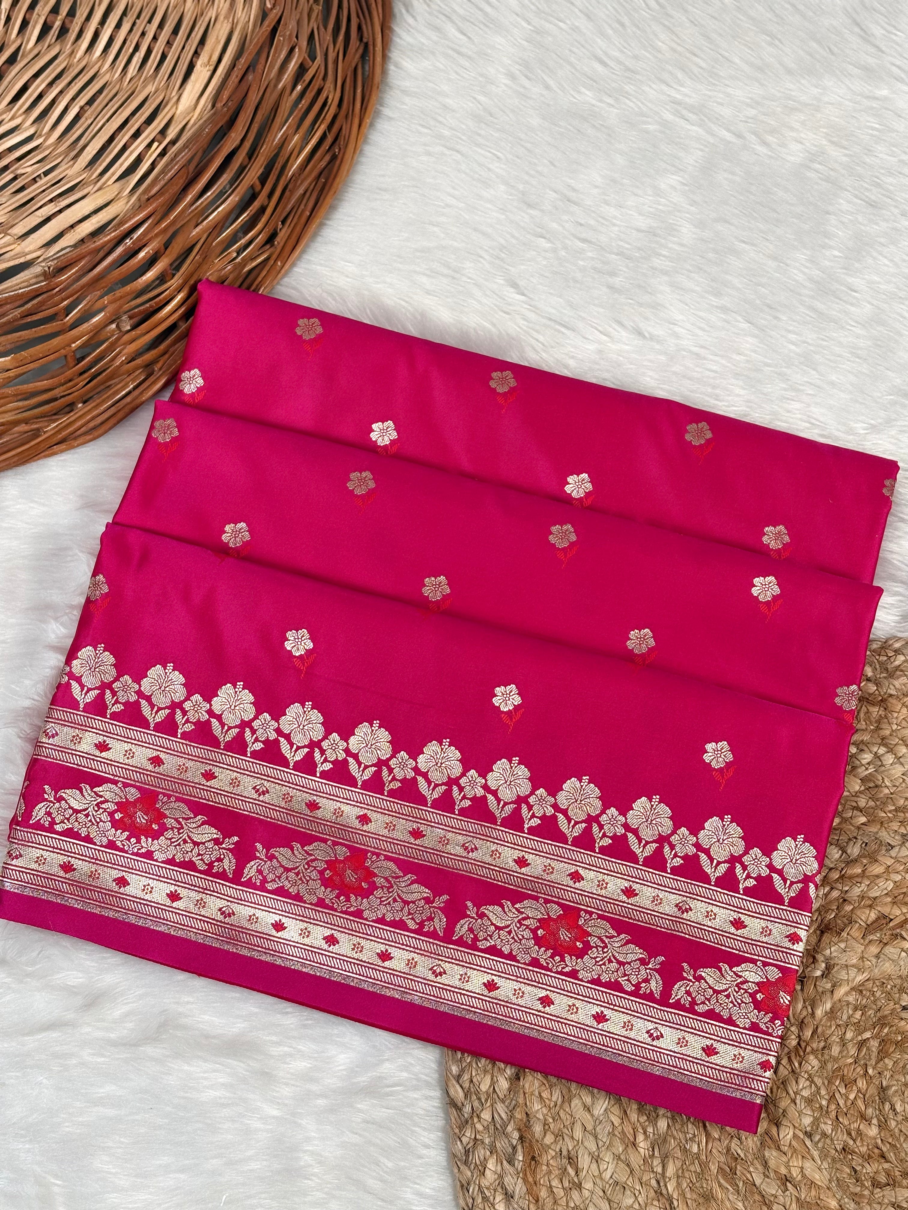 A folded magenta pink saree with intricate rose gold floral Banarasi zari weaving, displayed on a white faux fur surface next to a wicker basket.
