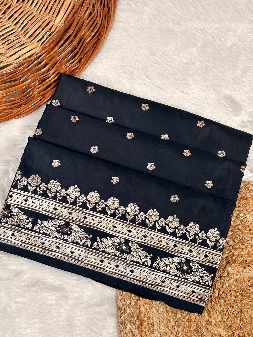 A folded black mashru katan saree with small scattered floral motifs and a wide decorative border featuring rose gold weaving, styled on a white faux-fur surface with woven props.