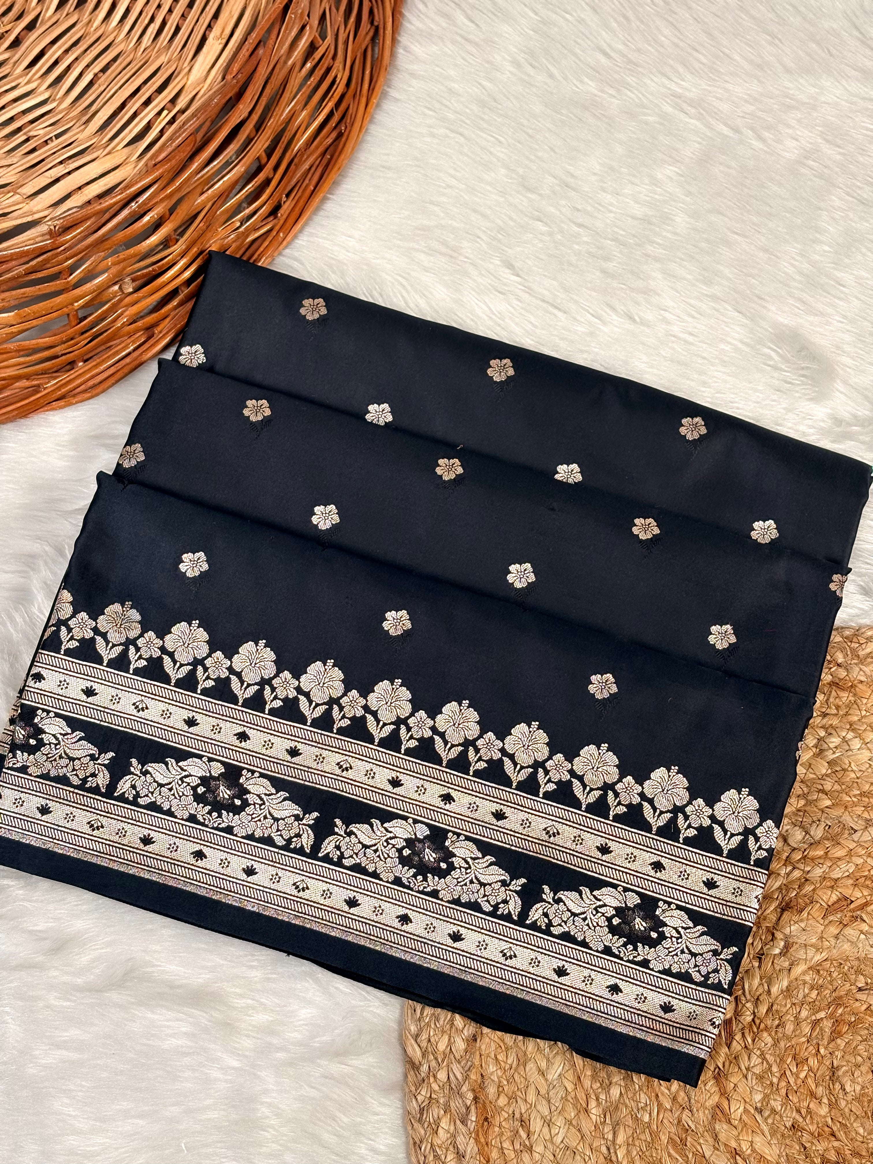 A folded black mashru katan saree with small scattered floral motifs and a wide decorative border featuring rose gold weaving, styled on a white faux-fur surface with woven props.