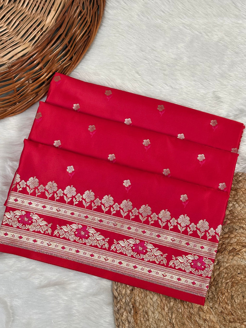 A folded red saree, featuring rose gold floral patterns and a wide decorative border, displayed on a white faux fur surface with a wicker basket as a prop.