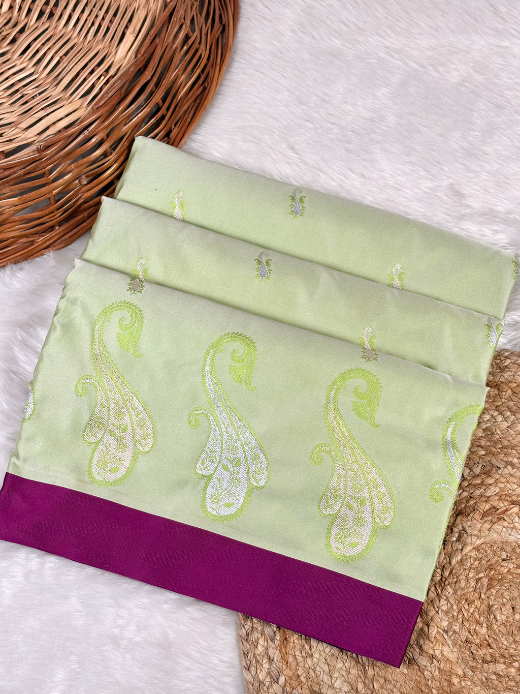 A folded light green saree, featuring paisley motifs and a contrasting magenta border, styled on a white faux fur surface with a woven wicker basket.