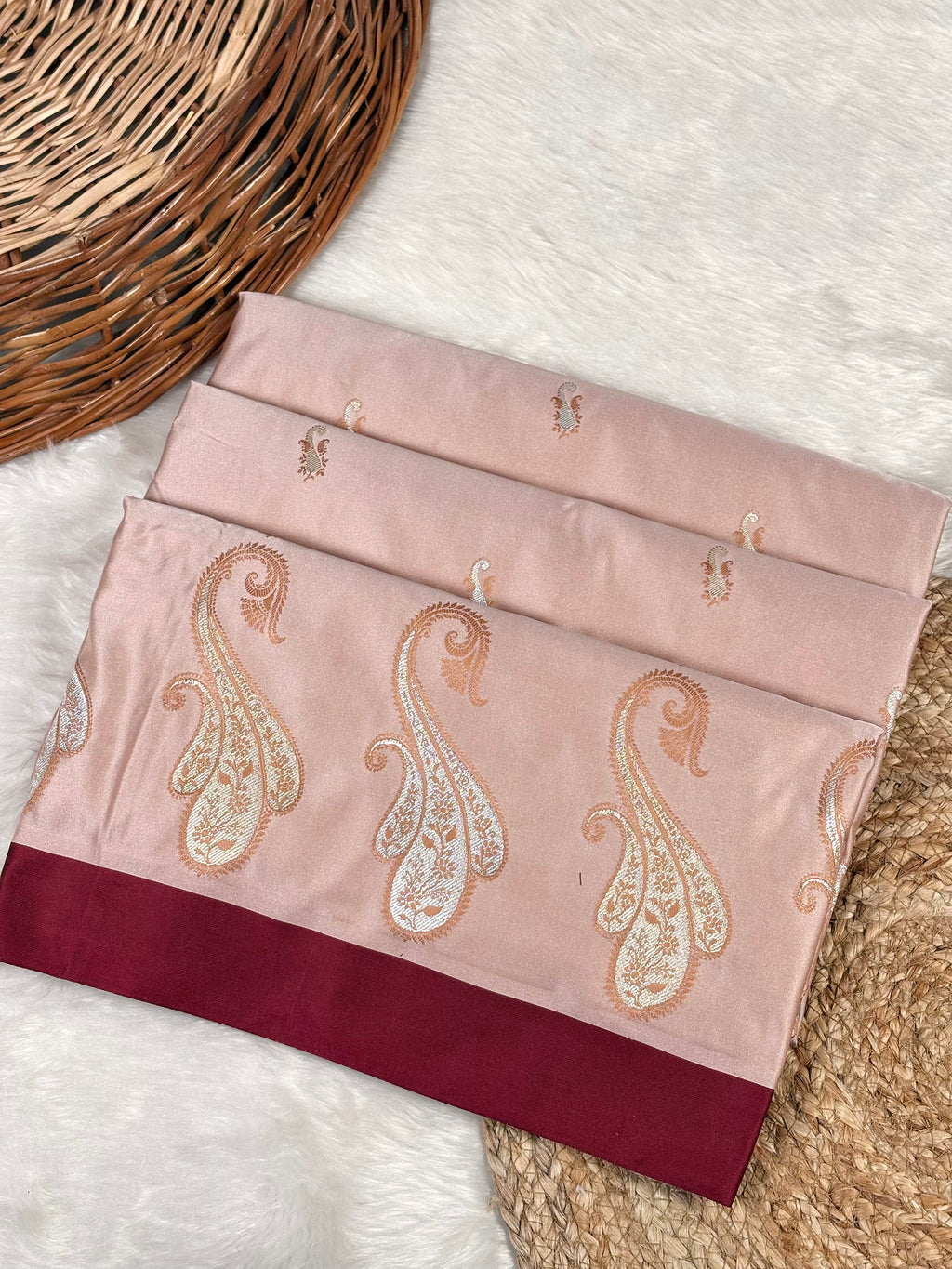 A folded dusty redfabric piece with metallic paisley weaving and a deep burgundy border, displayed on a white faux fur surface next to a woven wicker basket.