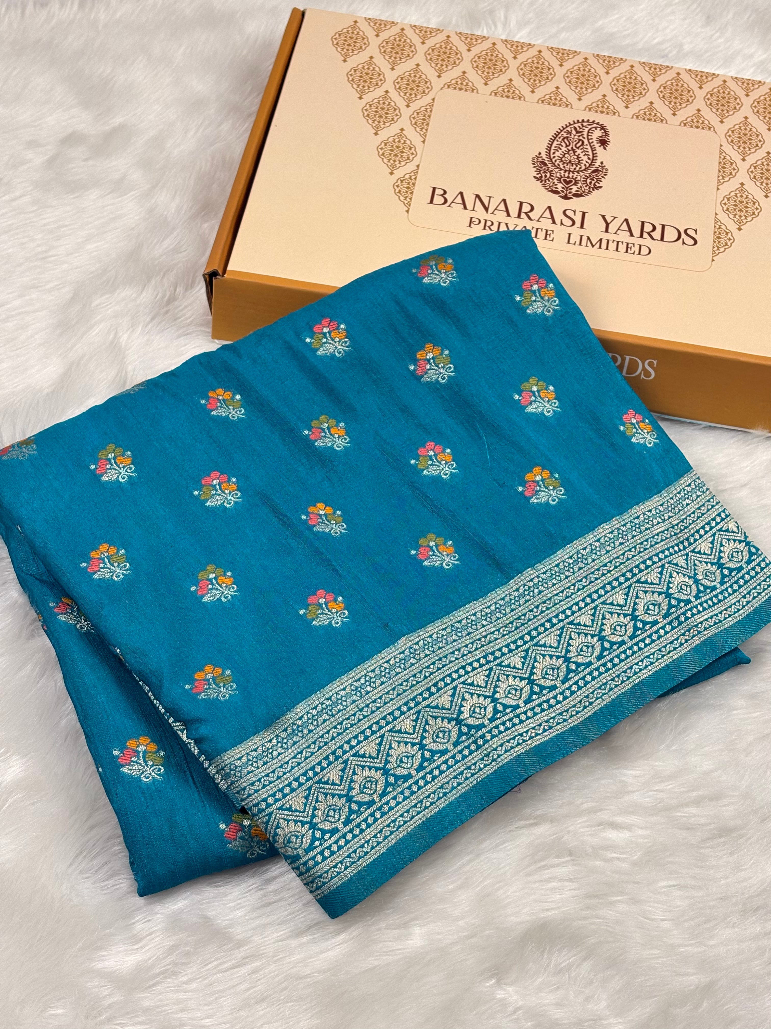 A folded teal blue saree with small multi-color floral motifs and a detailed white border, displayed next to a beige Banarasi Yards box on a white faux fur surface.