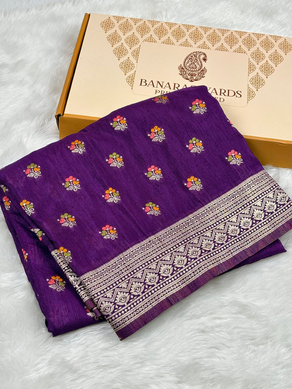 A folded purple Banarasi silk saree with colorful floral motifs and a detailed gold border, displayed next to its beige packaging box on a white faux fur surface.
