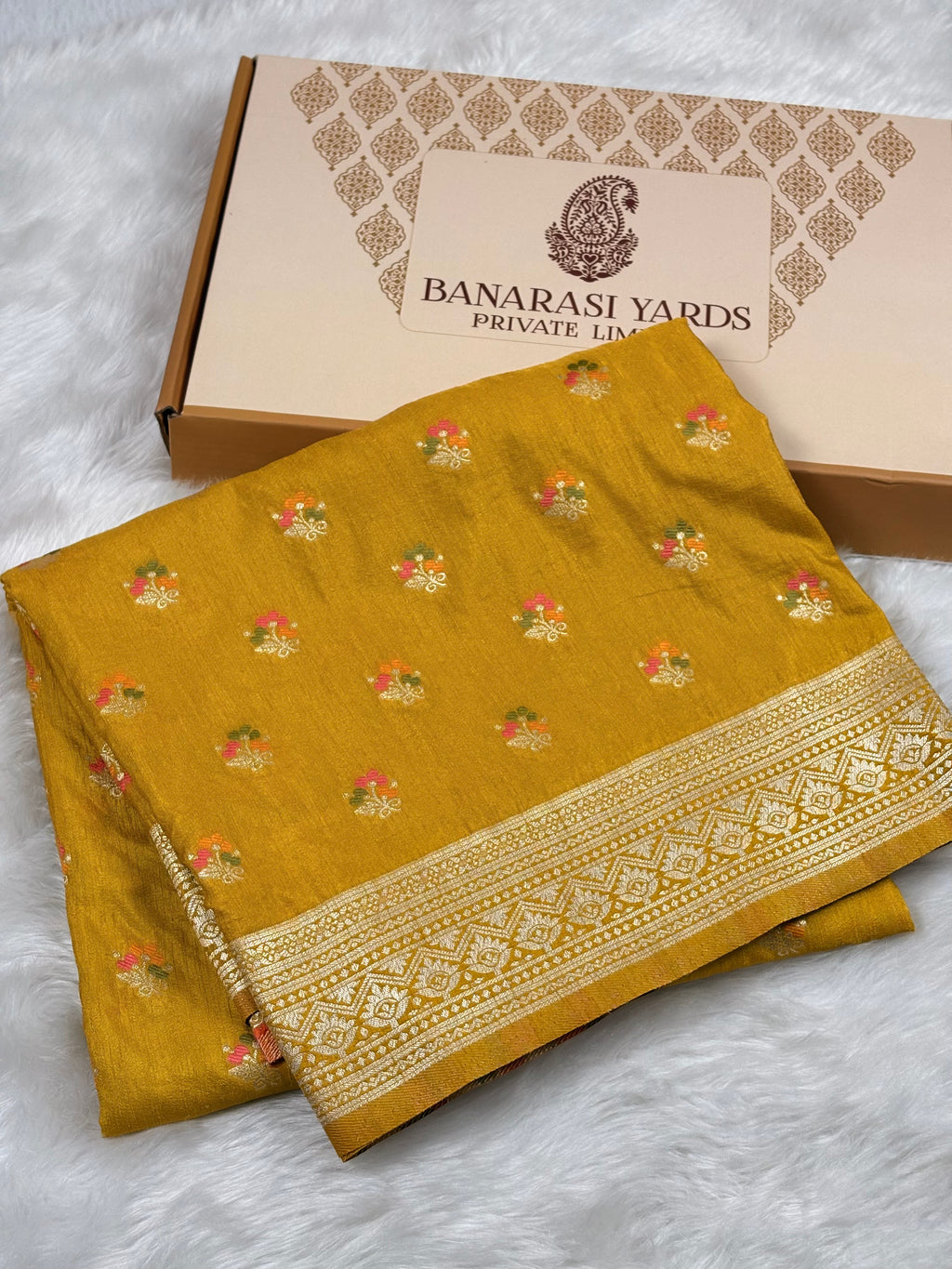 A folded mustard yellow Banarasi saree with small floral motifs and a decorative border, displayed in front of a branded Banarasi Yards box on a white faux-fur surface.
