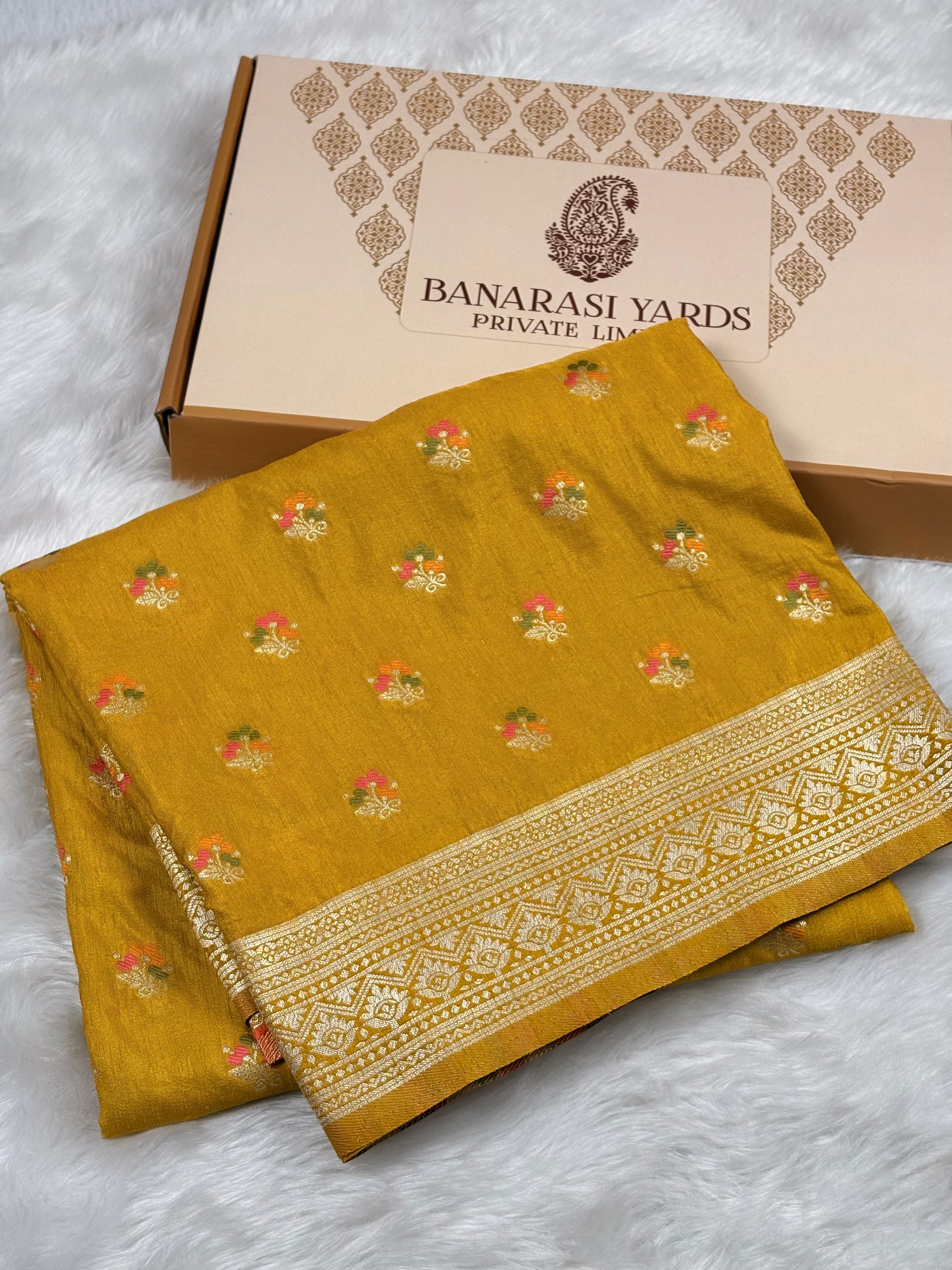 A folded mustard yellow Banarasi saree with small floral motifs and a decorative border, displayed in front of a branded Banarasi Yards box on a white faux-fur surface.