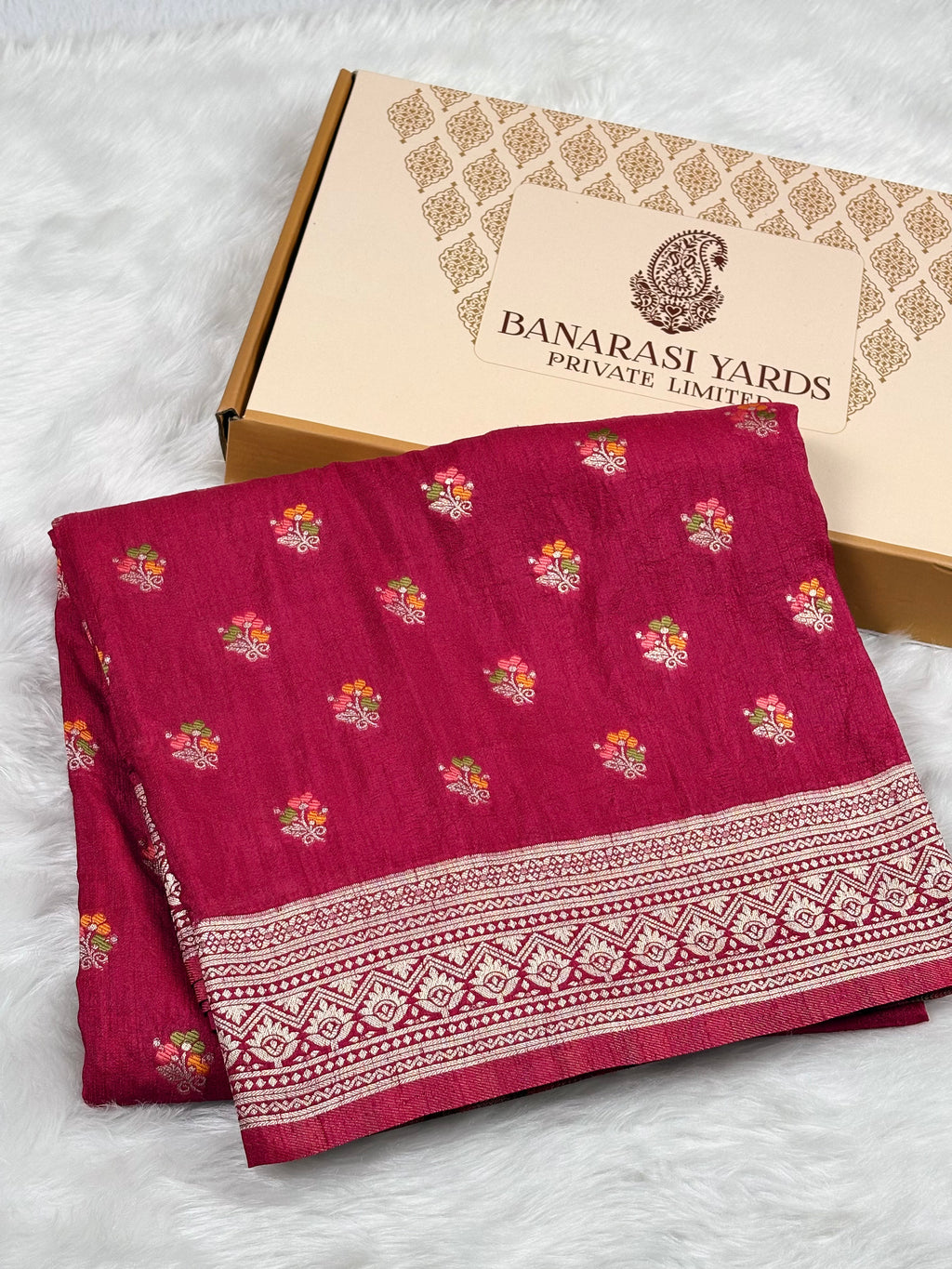 A folded deep pink Banarasi silk saree with colorful floral prints and a detailed zari border, displayed next to its beige packaging box on a white faux fur surface.