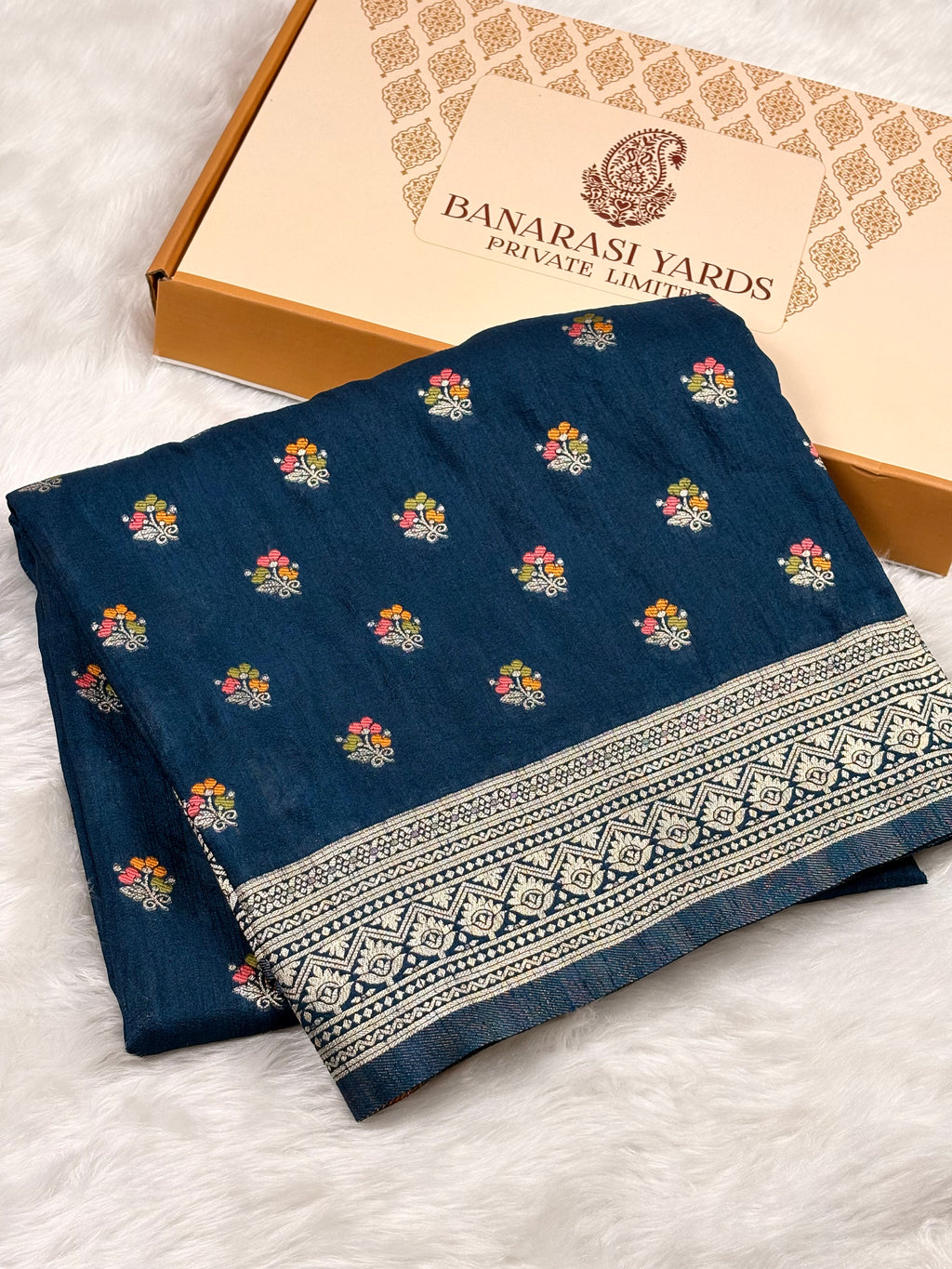 A folded navy blue Banarasi saree with small multi-color floral motifs and a wide zari geometric border, displayed next to a branded Banarasi Yards box on a white faux-fur surface.