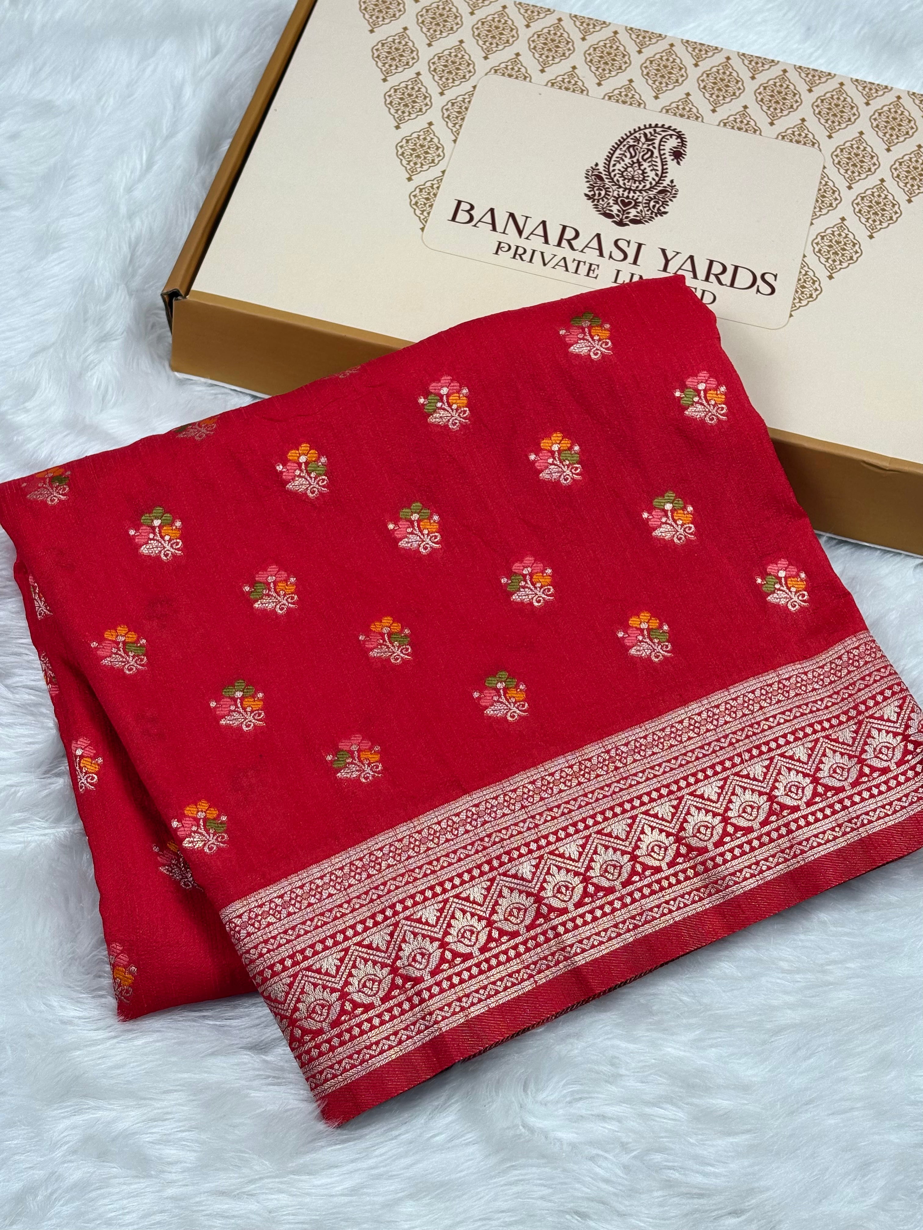 A folded red Banarasi saree with small floral motifs and a decorative border, displayed next to a beige Banarasi Yards box on a white faux-fur surface.