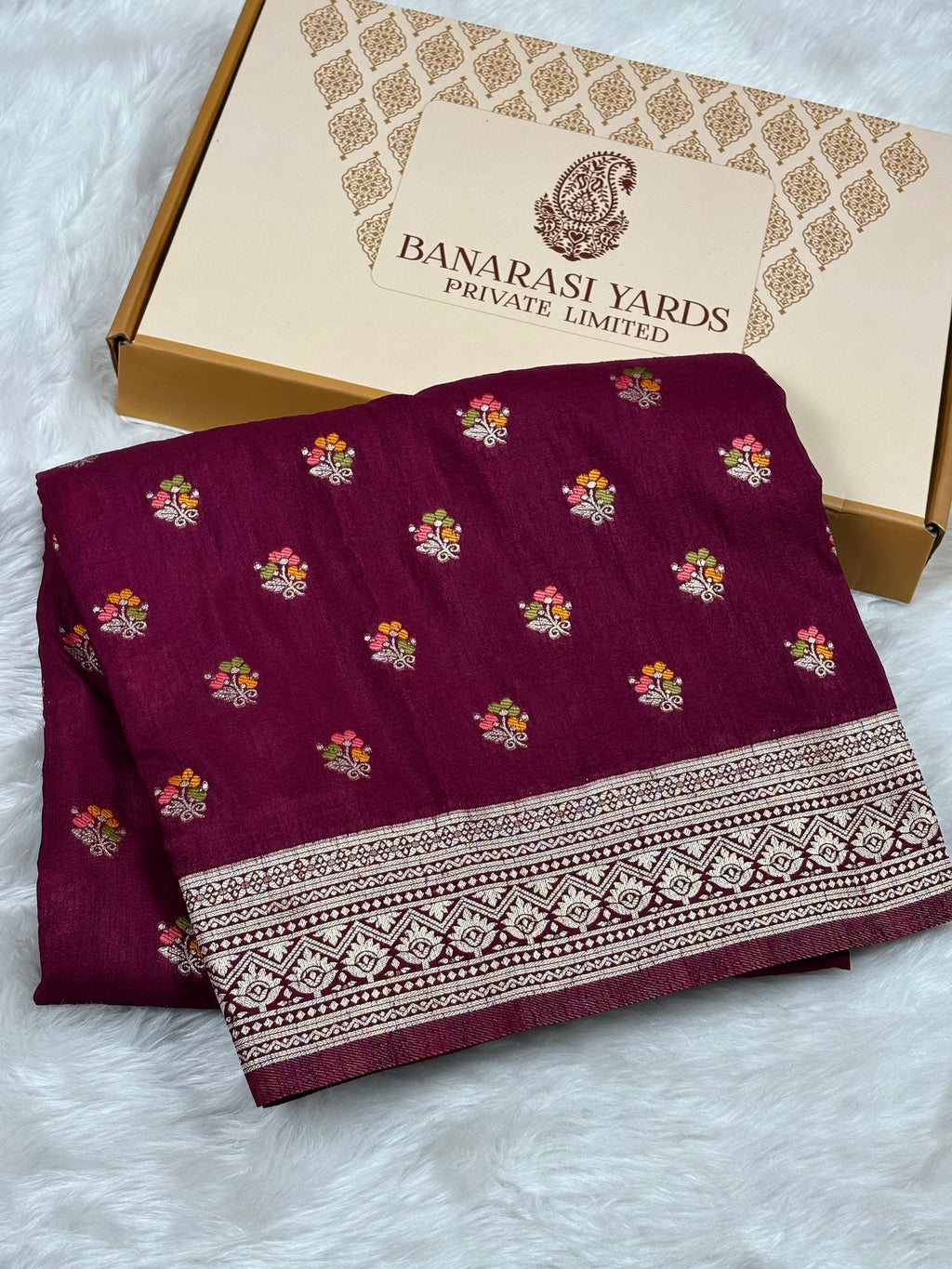 A folded deep magenta-purple fabric with small colorful floral motifs and a wide zari border, displayed next to a beige Banarasi Yards Private Limited box on a white faux-fur surface.
