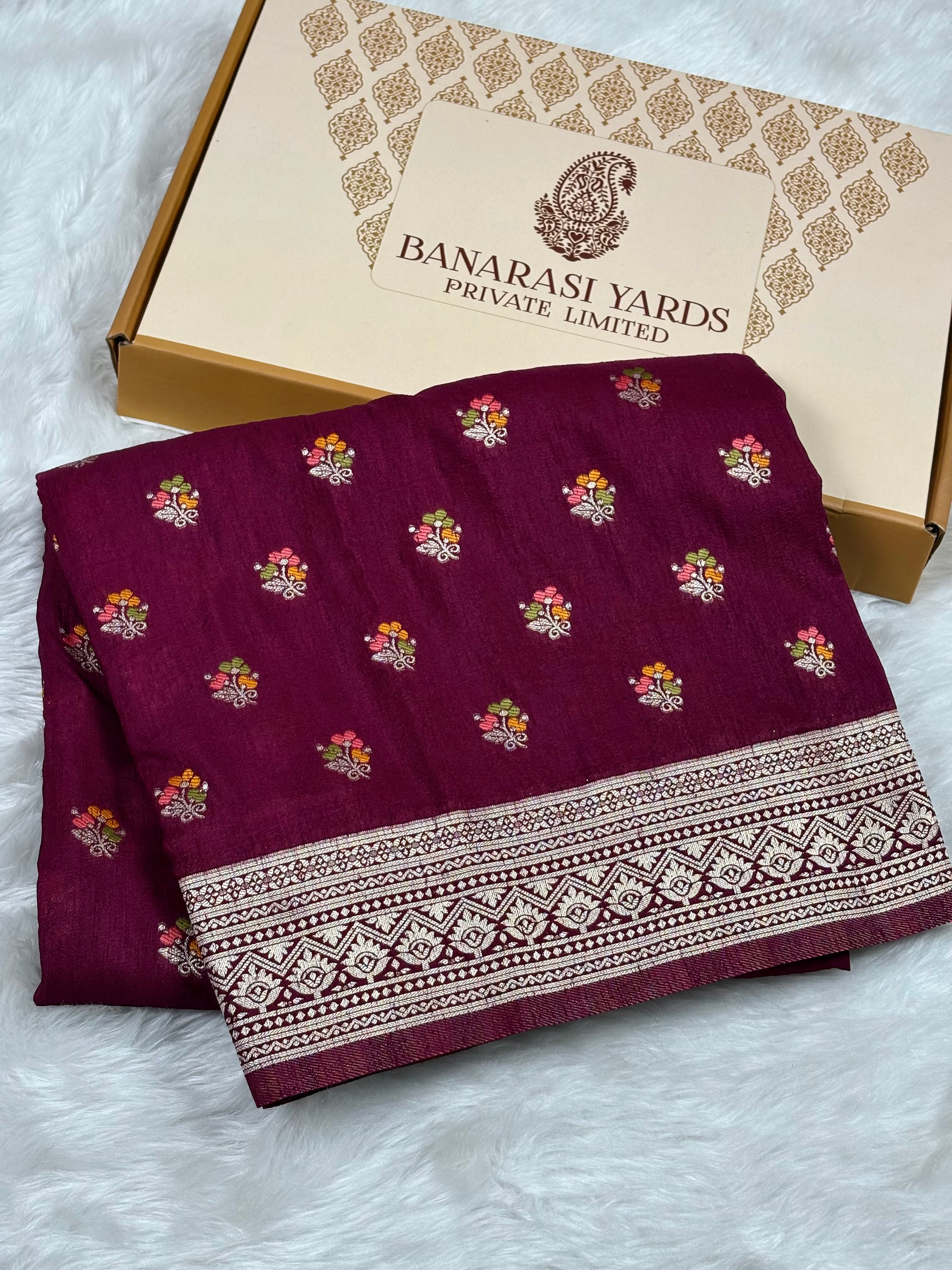 A folded deep magenta-purple fabric with small colorful floral motifs and a wide zari border, displayed next to a beige Banarasi Yards Private Limited box on a white faux-fur surface.