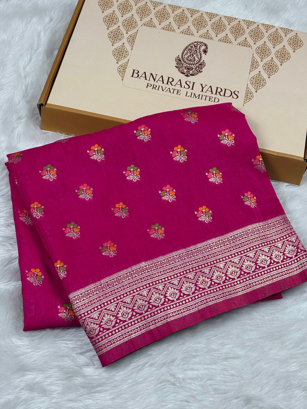 A folded magenta pink Banarasi line saree with small floral motifs and a detailed border, displayed next to its cardboard packaging box on a white faux fur surface.
