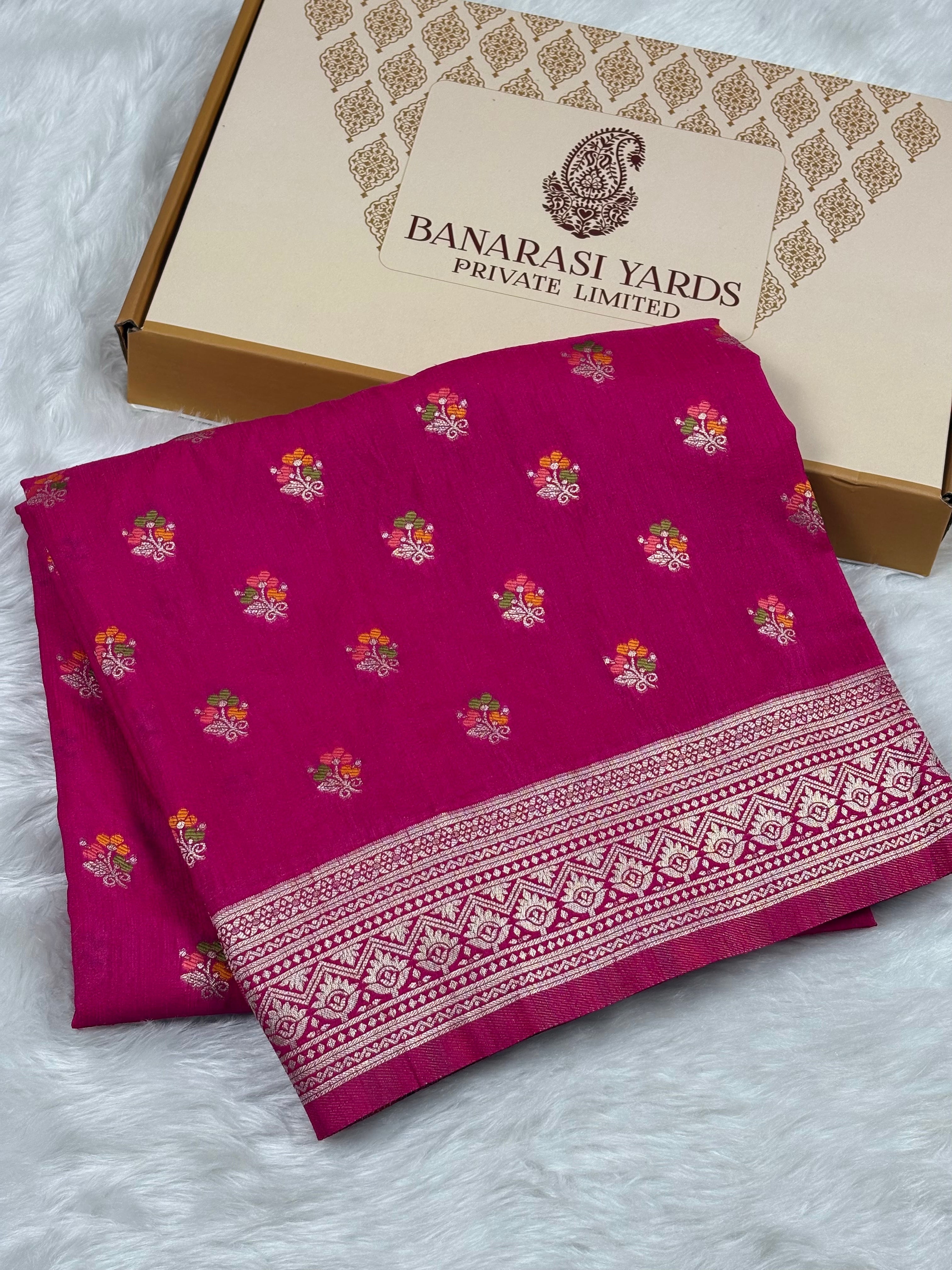 A folded magenta pink Banarasi line saree with small floral motifs and a detailed border, displayed next to its cardboard packaging box on a white faux fur surface.